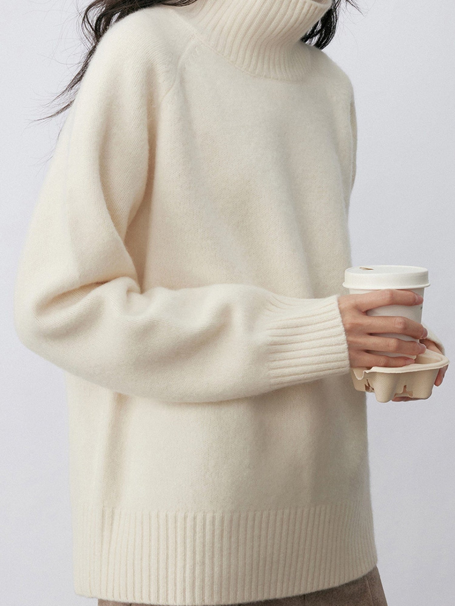 Woman wearing Arkin Studio minimalist turtleneck wool sweater in white and taupe pants, with a coffee cup in hands, standing sideways, on a white background