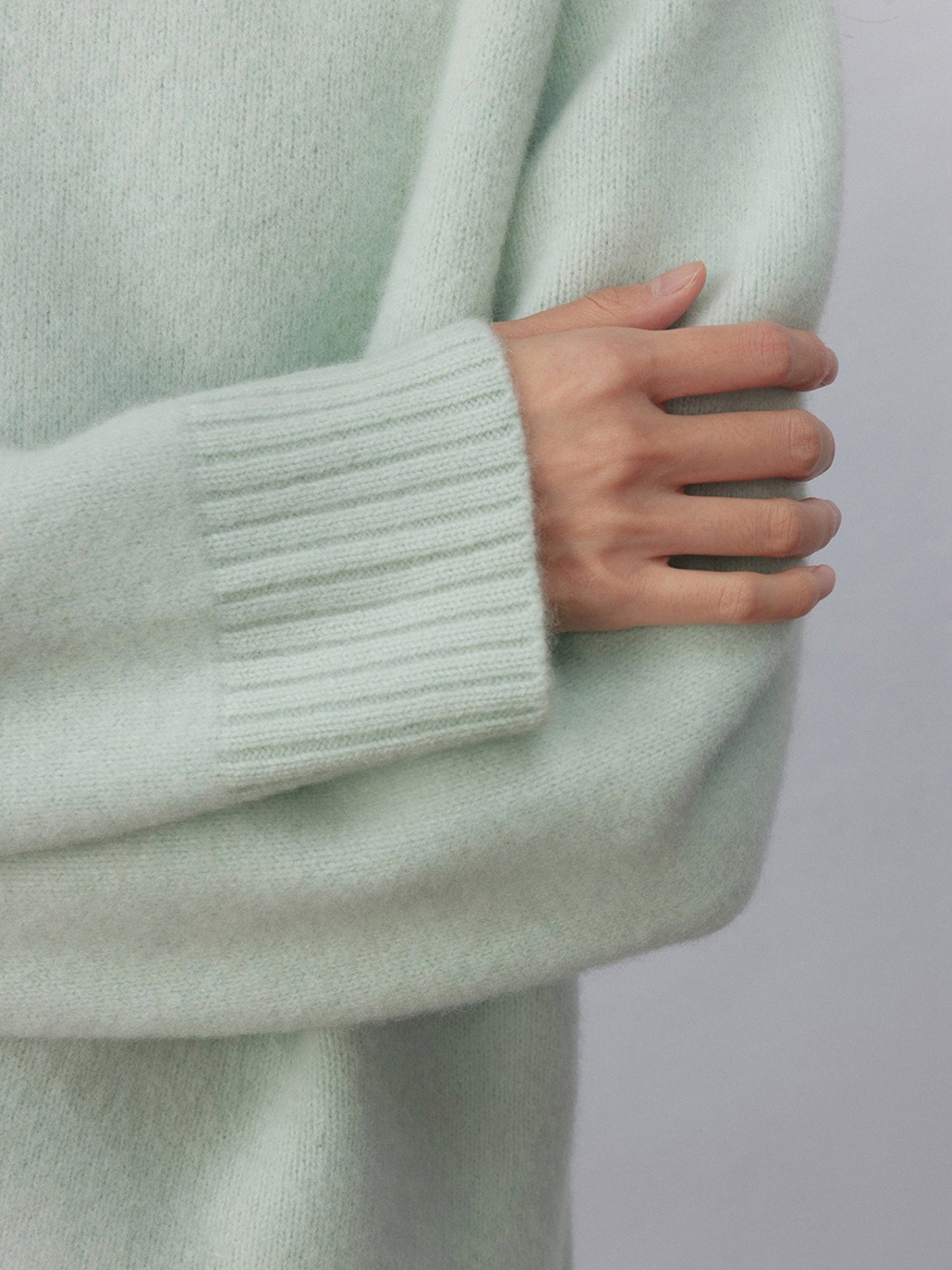 Close up of a woman wearing Arkin Studio minimalist turtleneck wool sweater in sage-green, arms crossing over her chest, standing facing the camera, on a white background