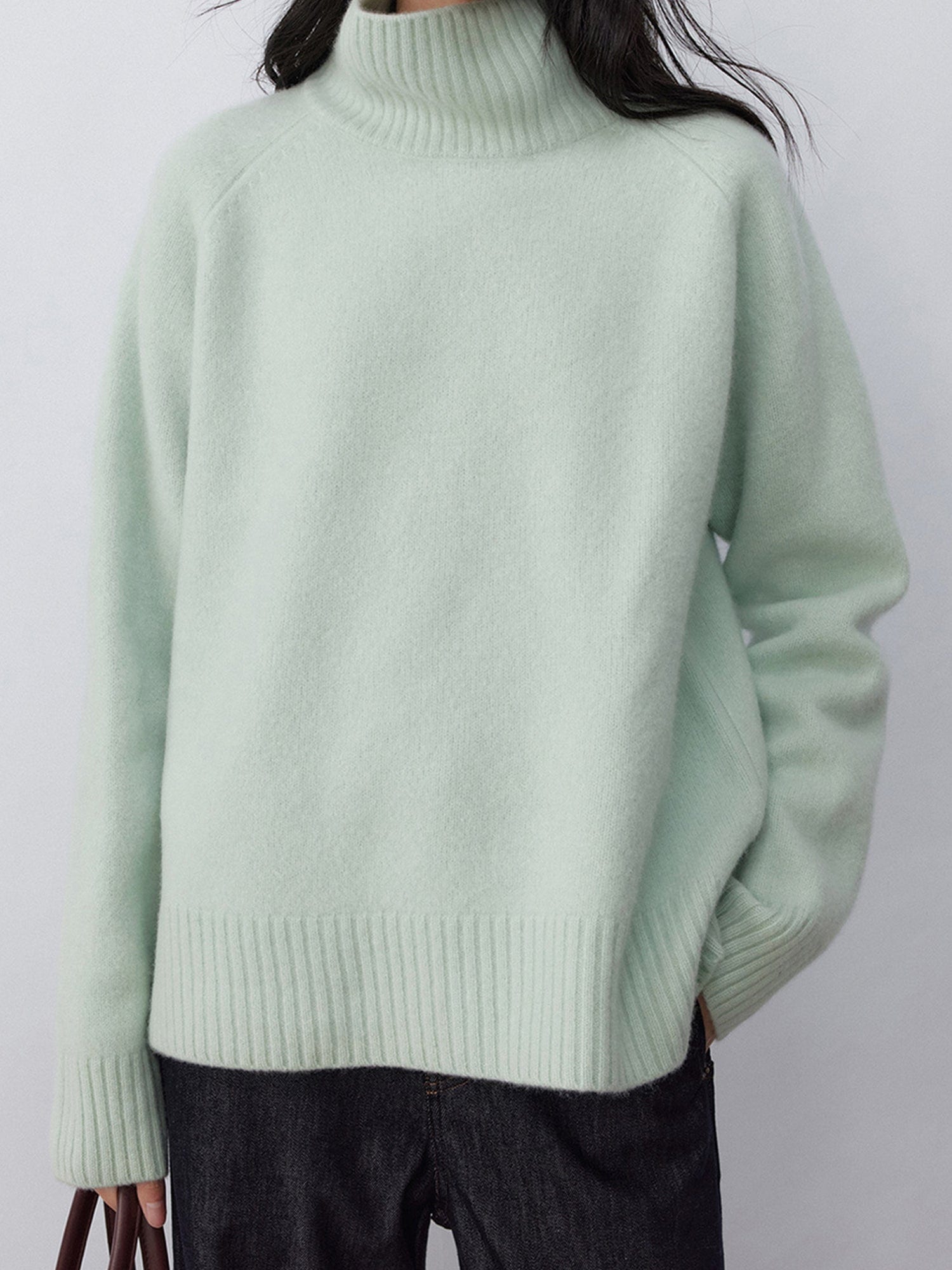 Woman wearing Arkin Studio minimalist turtleneck wool sweater in sage-green and dark jeans, holding a bag, standing facing the camera, on a white background