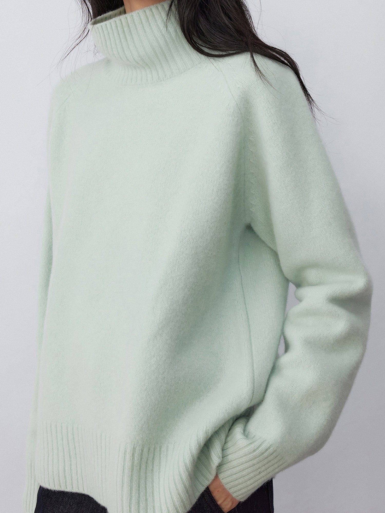 Woman wearing Arkin Studio minimalist turtleneck wool sweater in sage-green and dark jeans, standing sideways, on a white background