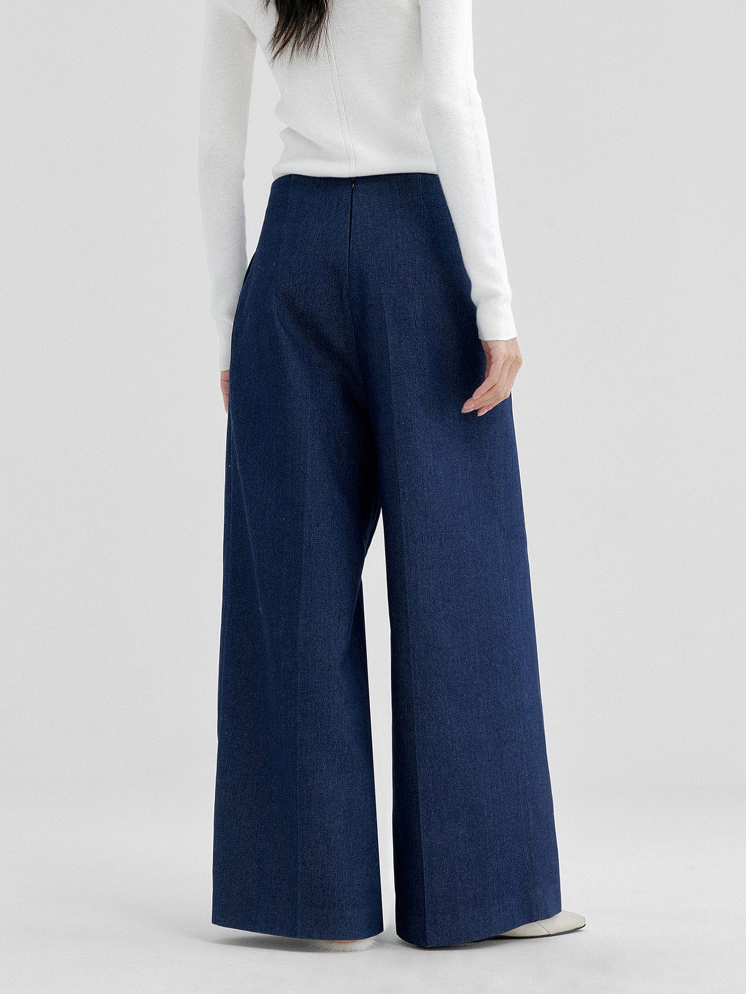 Woman wearing Arkin Studio no waistband wide leg jeans in navy and white a long sleeve top, with hands down at her sides, standing facing a white wall, on a white background
