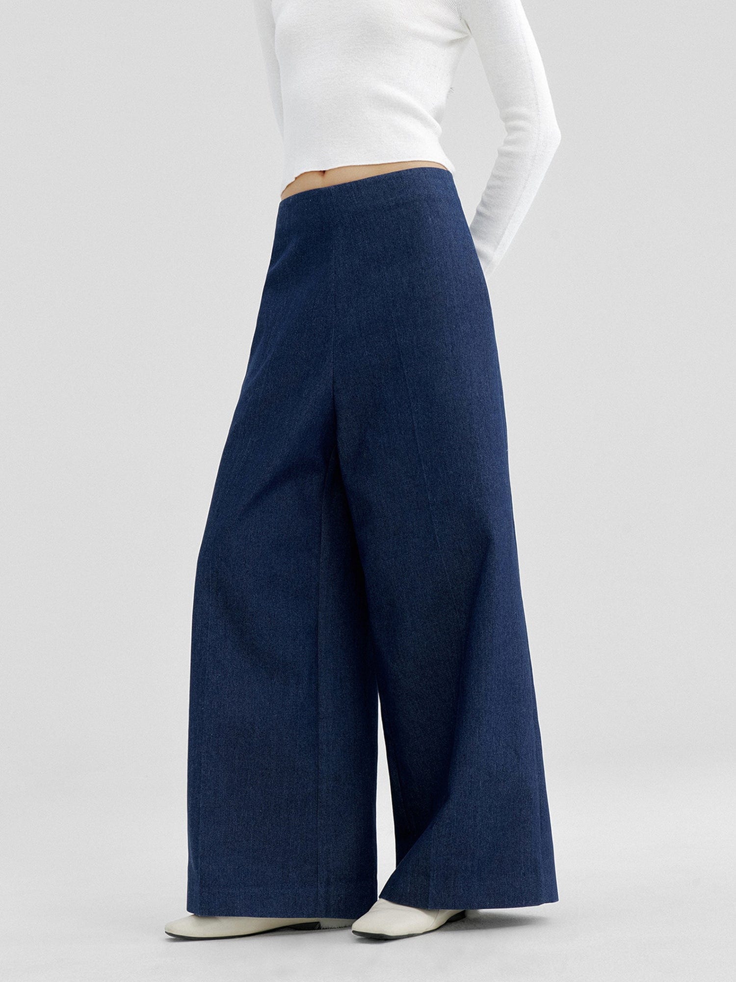 Woman wearing Arkin Studio no waistband wide leg jeans in navy and white a long sleeve top, with hands behind her back, standing sideways, on a white background