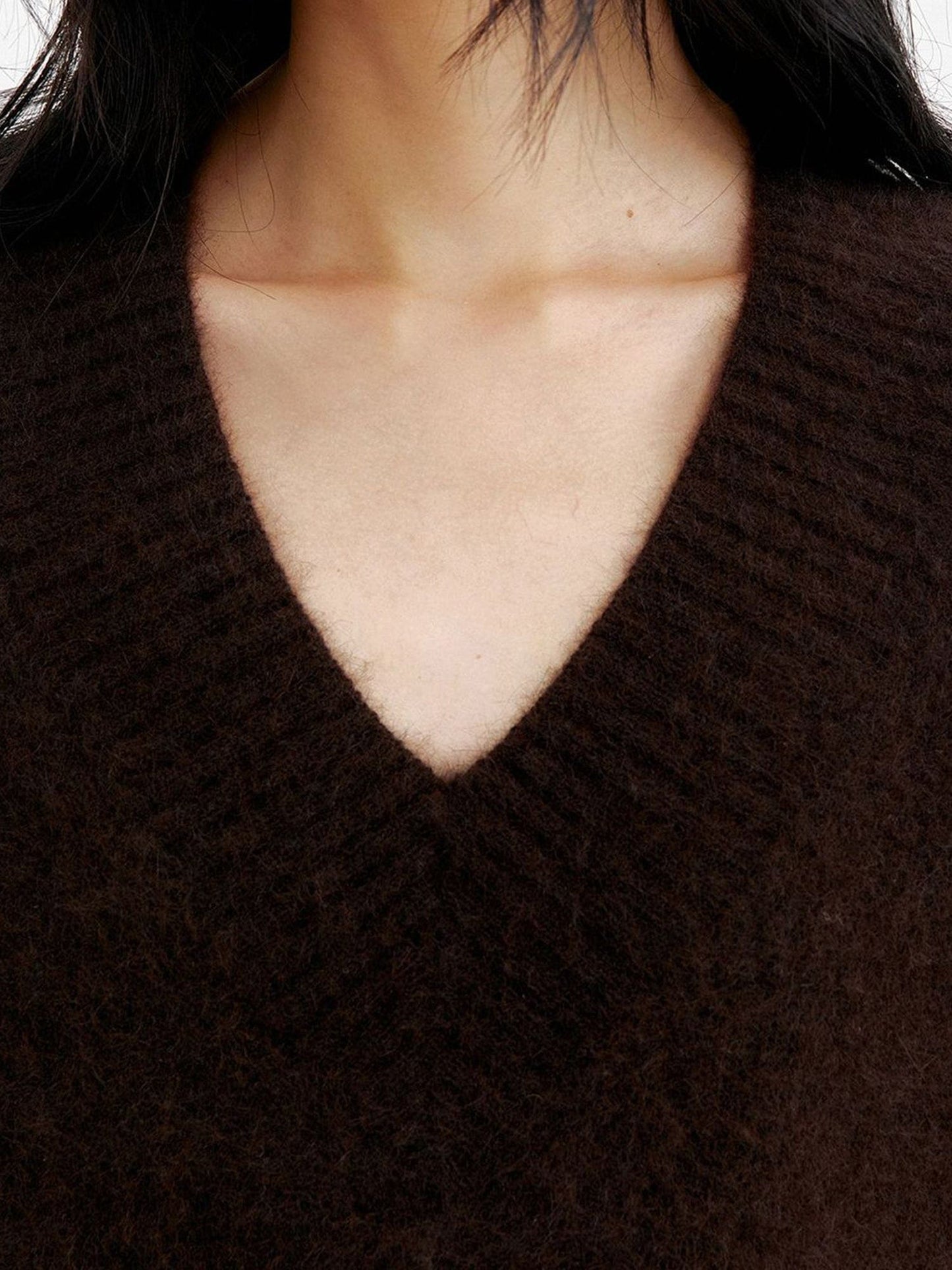 Close-up of the v-neck area of a woman wearing Arkin Studio oversized v-neck fuzzy sweater in dark-brown, facing the camera, on a white background