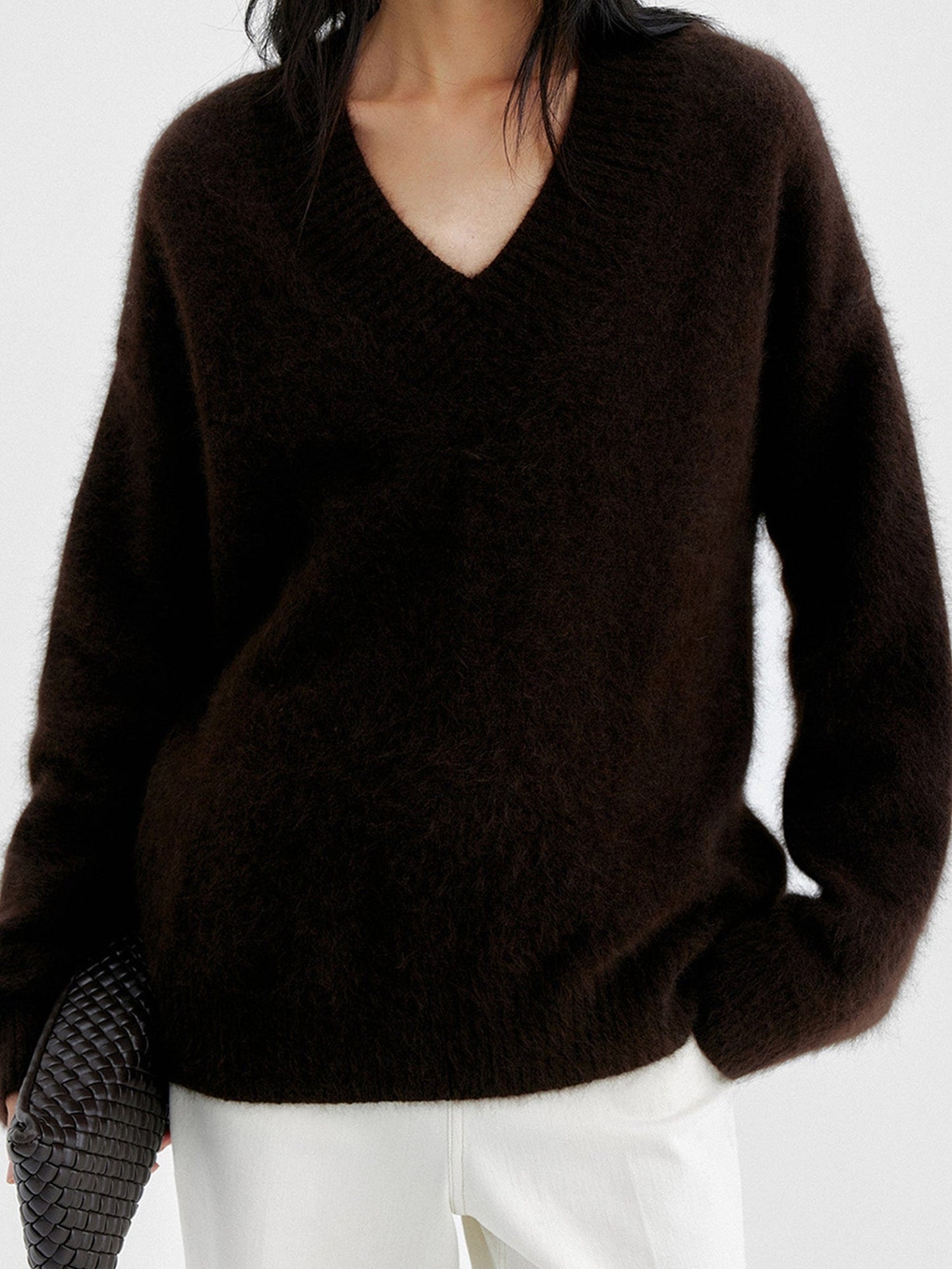 Woman wearing Arkin Studio oversized v-neck fuzzy sweater in dark-brown and matching white pants, holding a black bag, standing facing the camera, on a white background