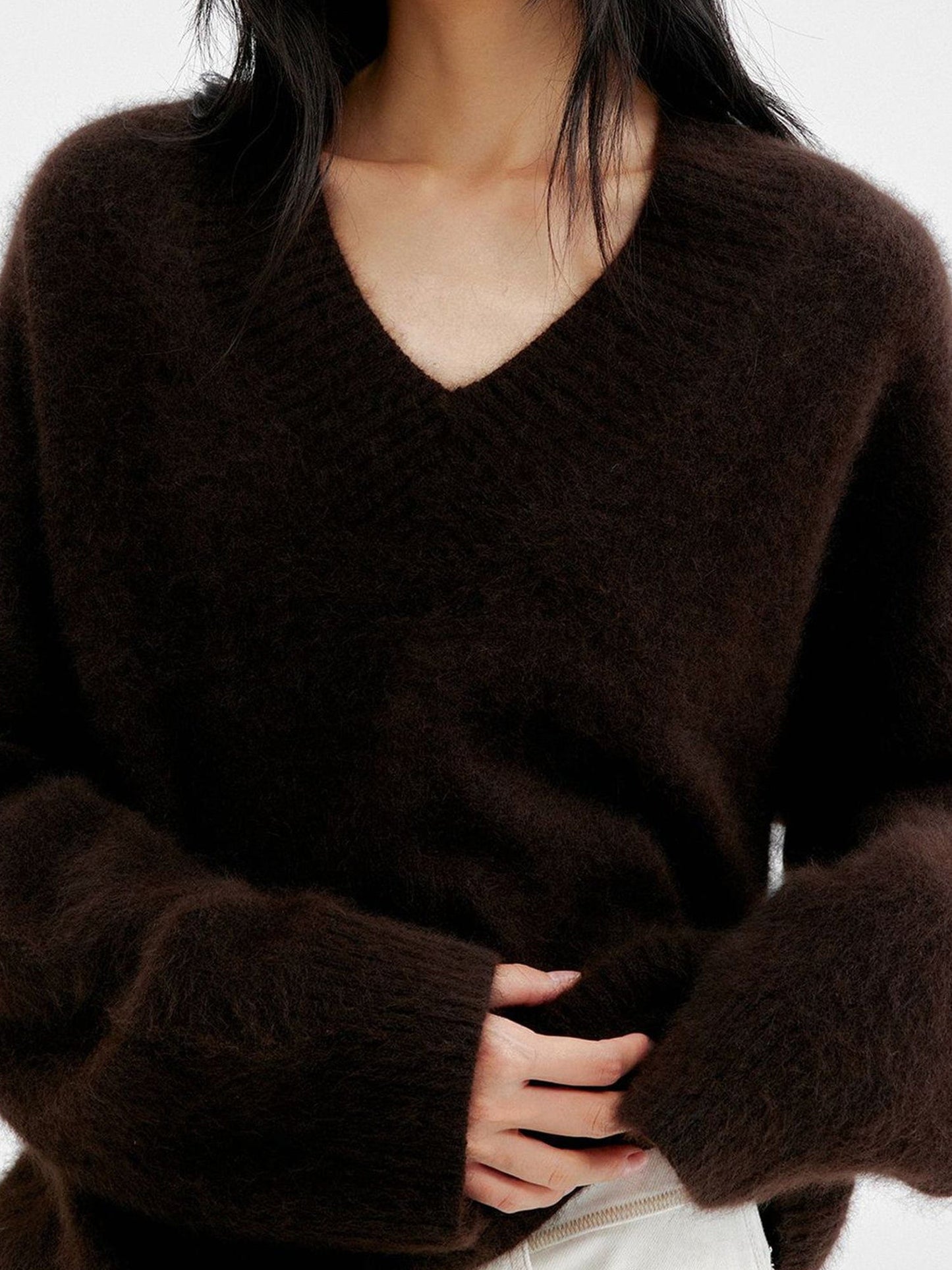 Woman wearing Arkin Studio oversized v-neck fuzzy sweater in dark-brown and matching white pants, hands touching the waist, standing facing the camera, on a white background