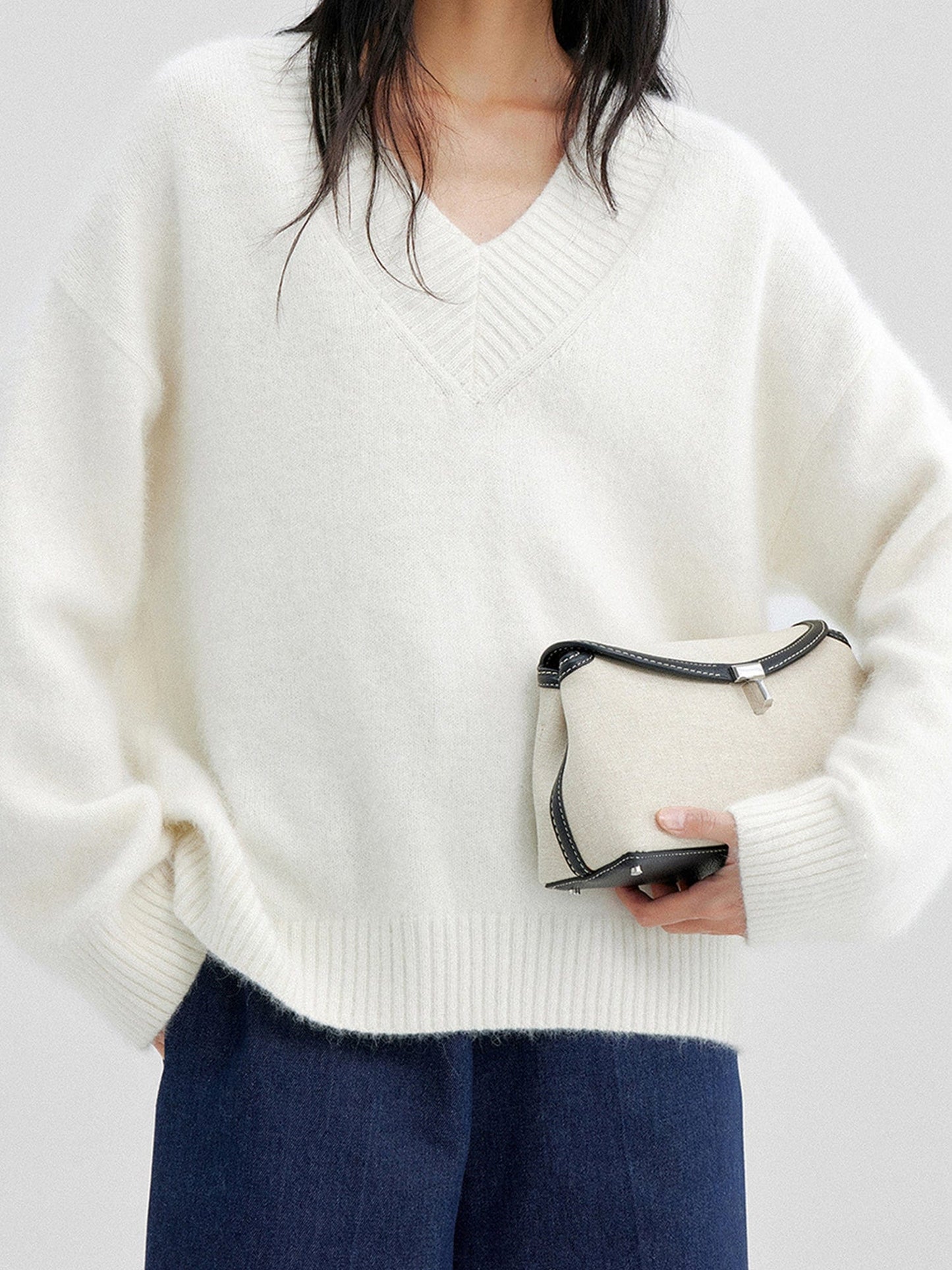 Woman wearing Arkin Studio oversized v-neck fuzzy sweater in white and matching navy pants, holding a white bag, standing facing the camera, on a white background