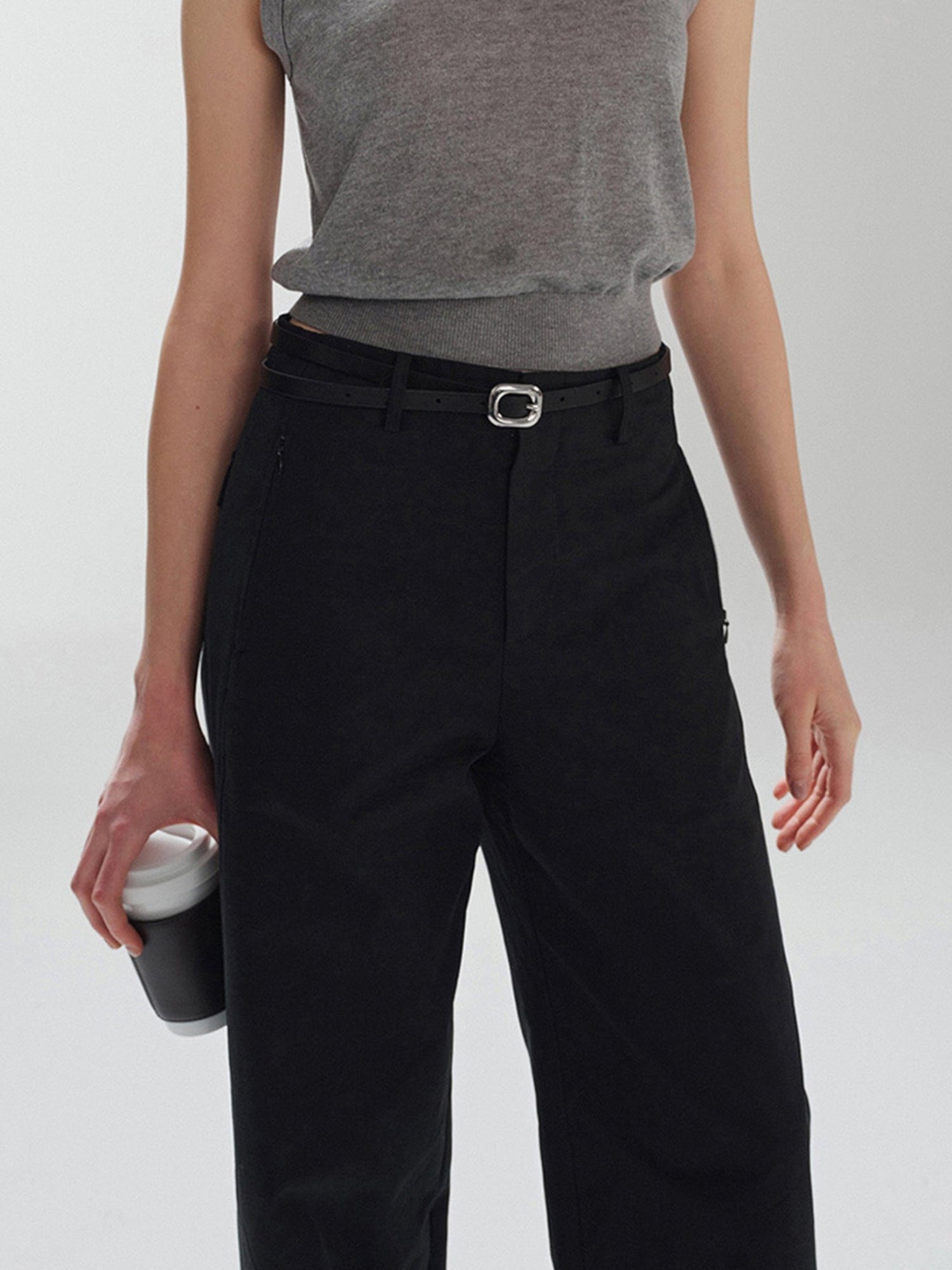 Woman wearing Arkin Studio pear friendly curved leg pants in black and a gray top, with coffee in right hand, standing facing the camera, on a white background