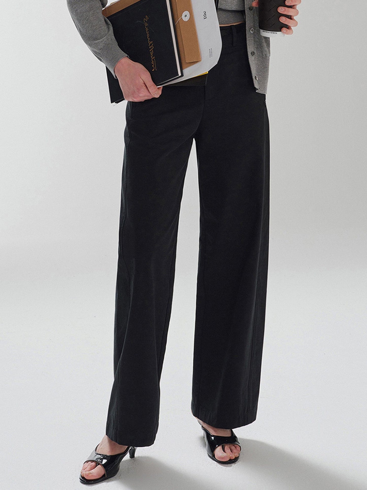 Woman wearing Arkin Studio pear friendly curved leg pants in black and a gray top, with coffee and documents in hands, standing facing the camera, on a white background