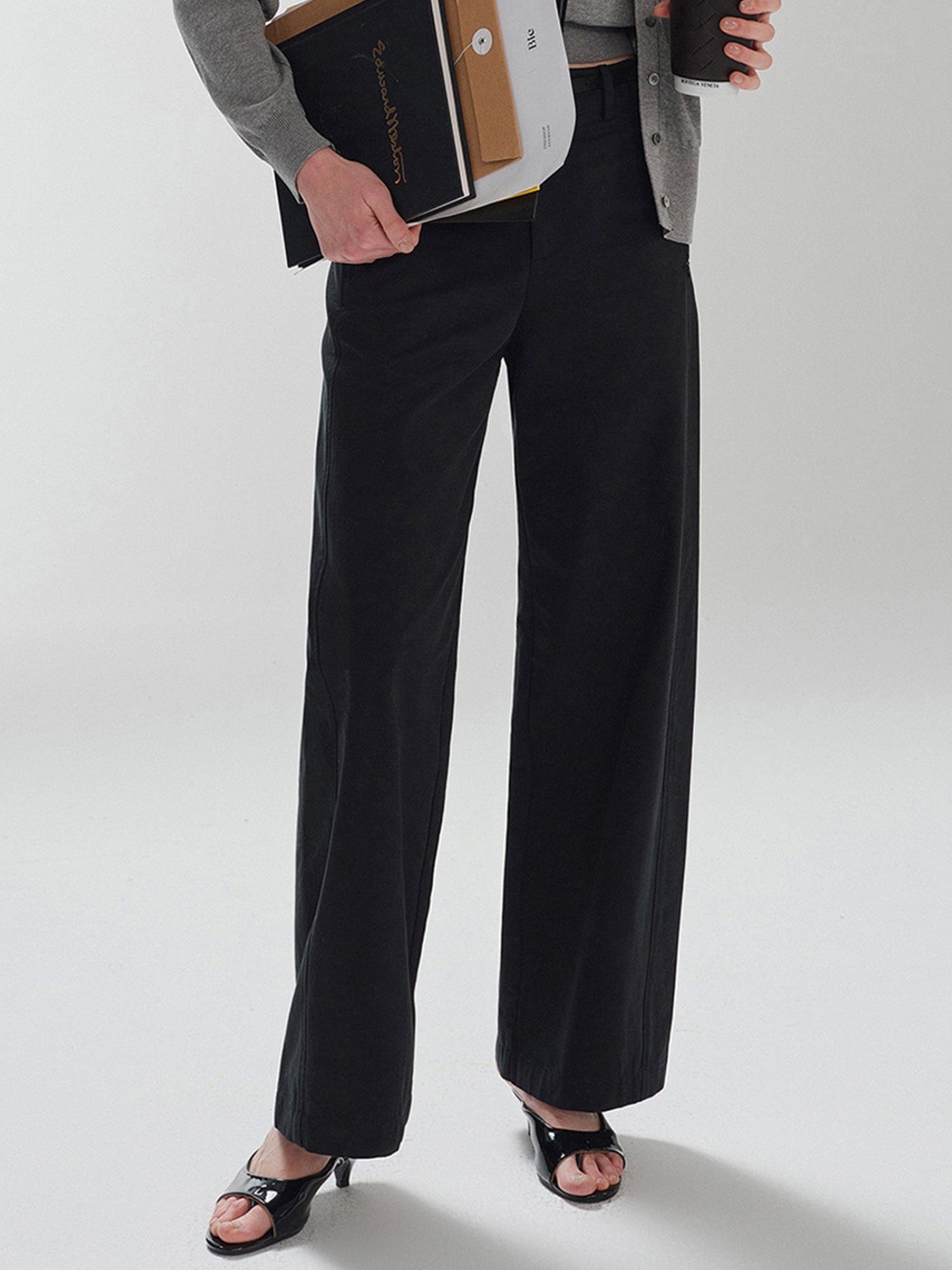 Woman wearing Arkin Studio pear friendly curved leg pants in black and a gray top, with coffee and documents in hands, standing facing the camera, on a white background