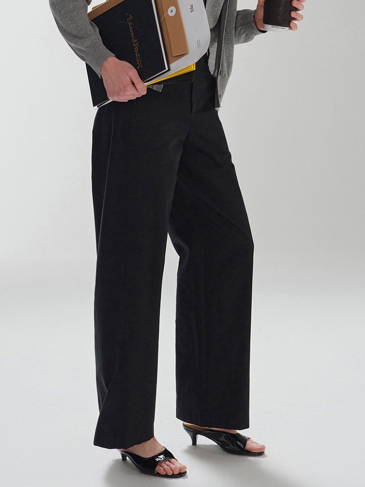 Woman wearing Arkin Studio pear friendly curved leg pants in black and a gray top, with coffee and documents in hands, standing sideways, on a white background