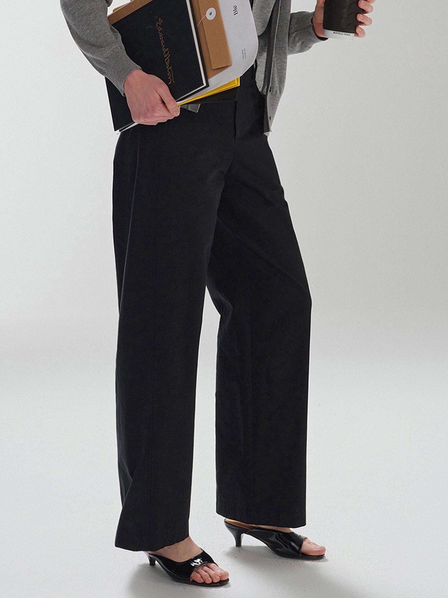 Woman wearing Arkin Studio pear friendly curved leg pants in black and a gray top, with coffee and documents in hands, standing sideways, on a white background