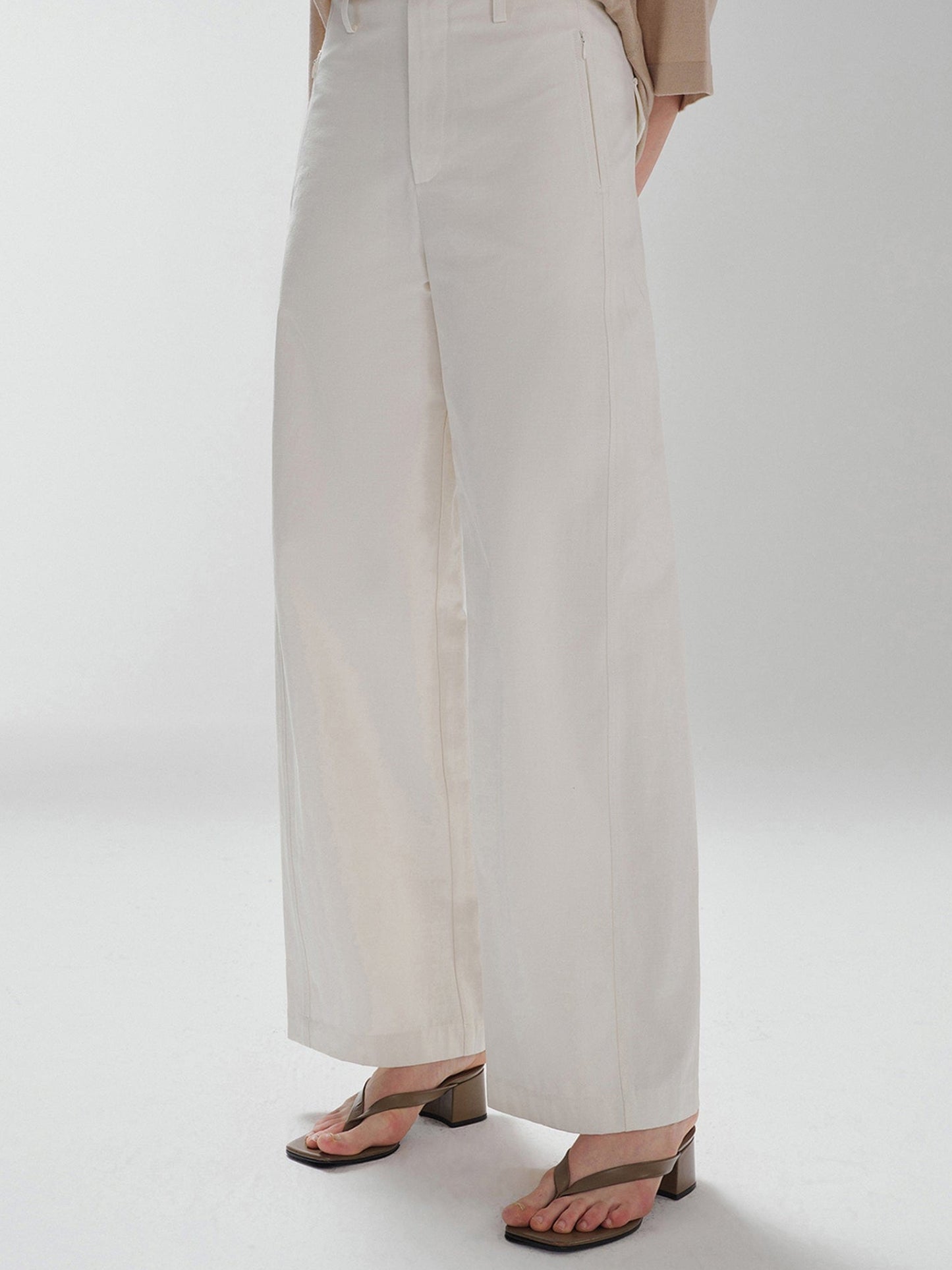 Woman wearing Arkin Studio pear friendly curved leg pants in ivory and a brown top, with hands clasped behind her back, body slightly turned to the right, standing facing the camera, on a white background