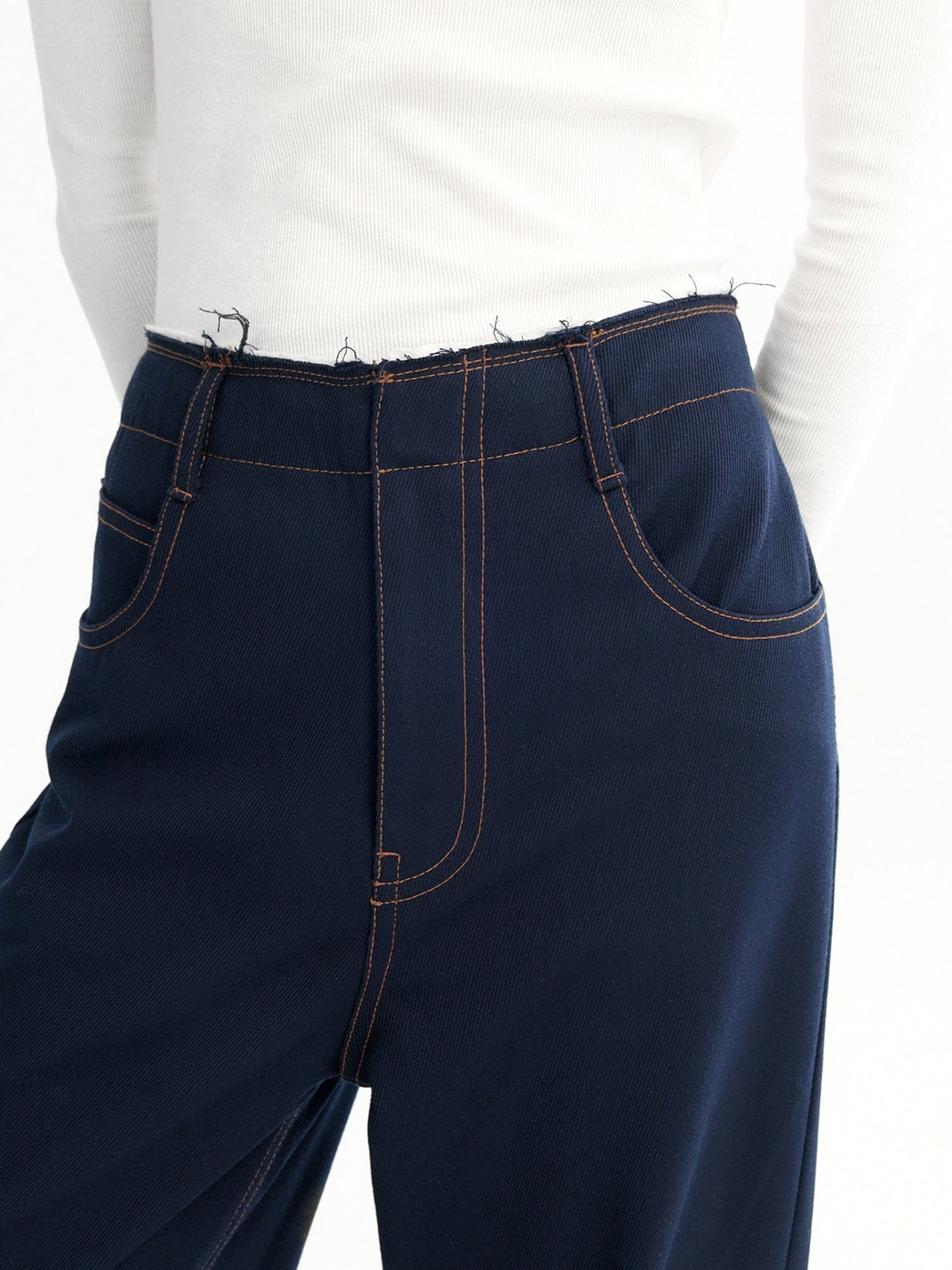 Close-up of a woman's waist and abdomen wearing Arkin Studio dark navy raw edge contrast stitch trousers and a white long-sleeve tee, with hands behind her back, standing facing the camera, focusing on the waistband and waist design, on a white background