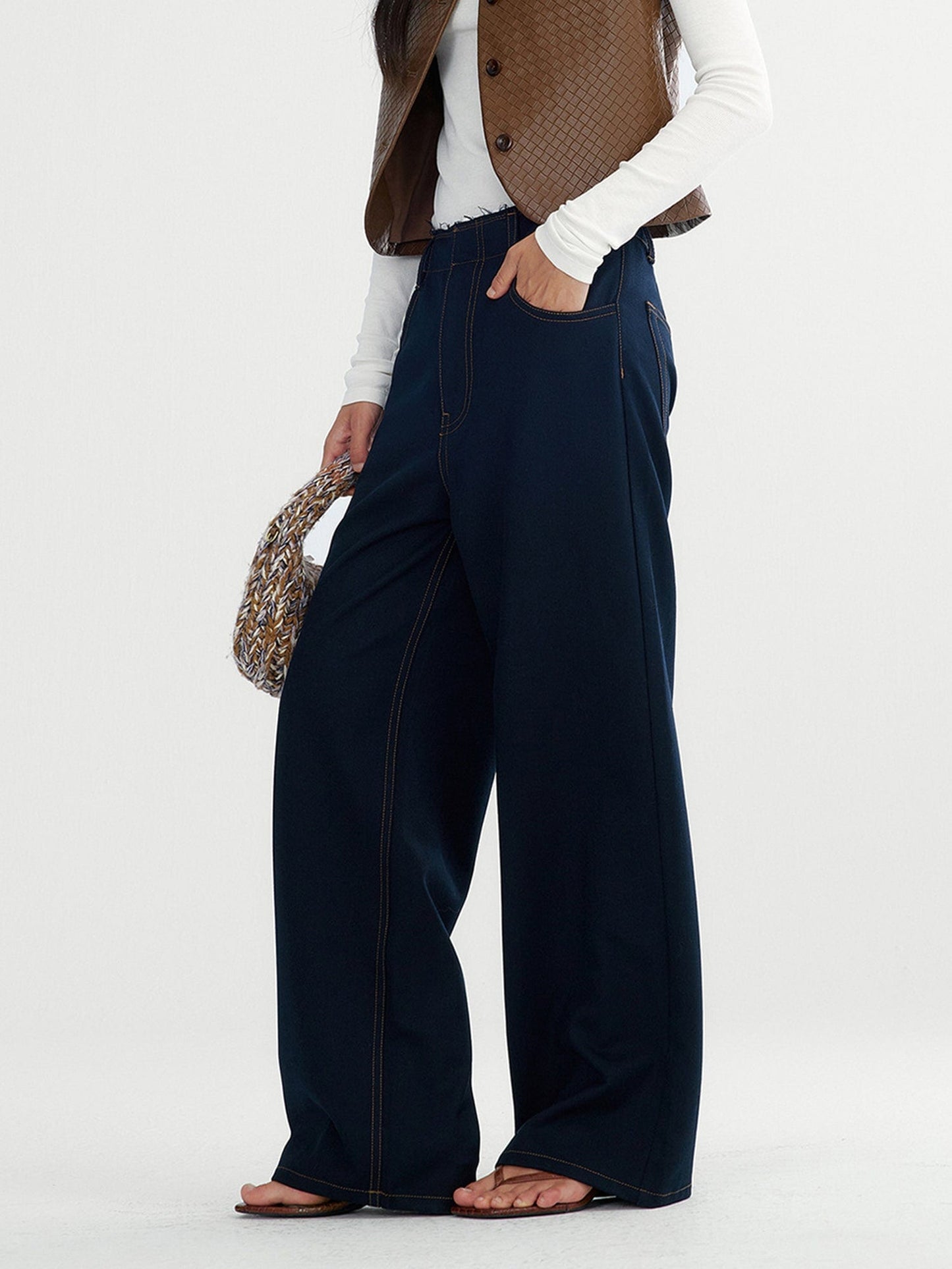 Woman wearing Arkin Studio raw edge contrast stitch trousers in dark navy, a white long-sleeve tee, and a brown vest, with one hand in her pocket and another holding a woven bag, standing sideways, on a white background