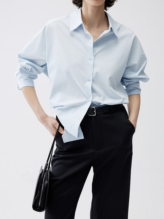 Woman wearing Arkin Studio relaxed blue tencel cotton blouse in light blue and black pants, standing facing the camera, on a white background