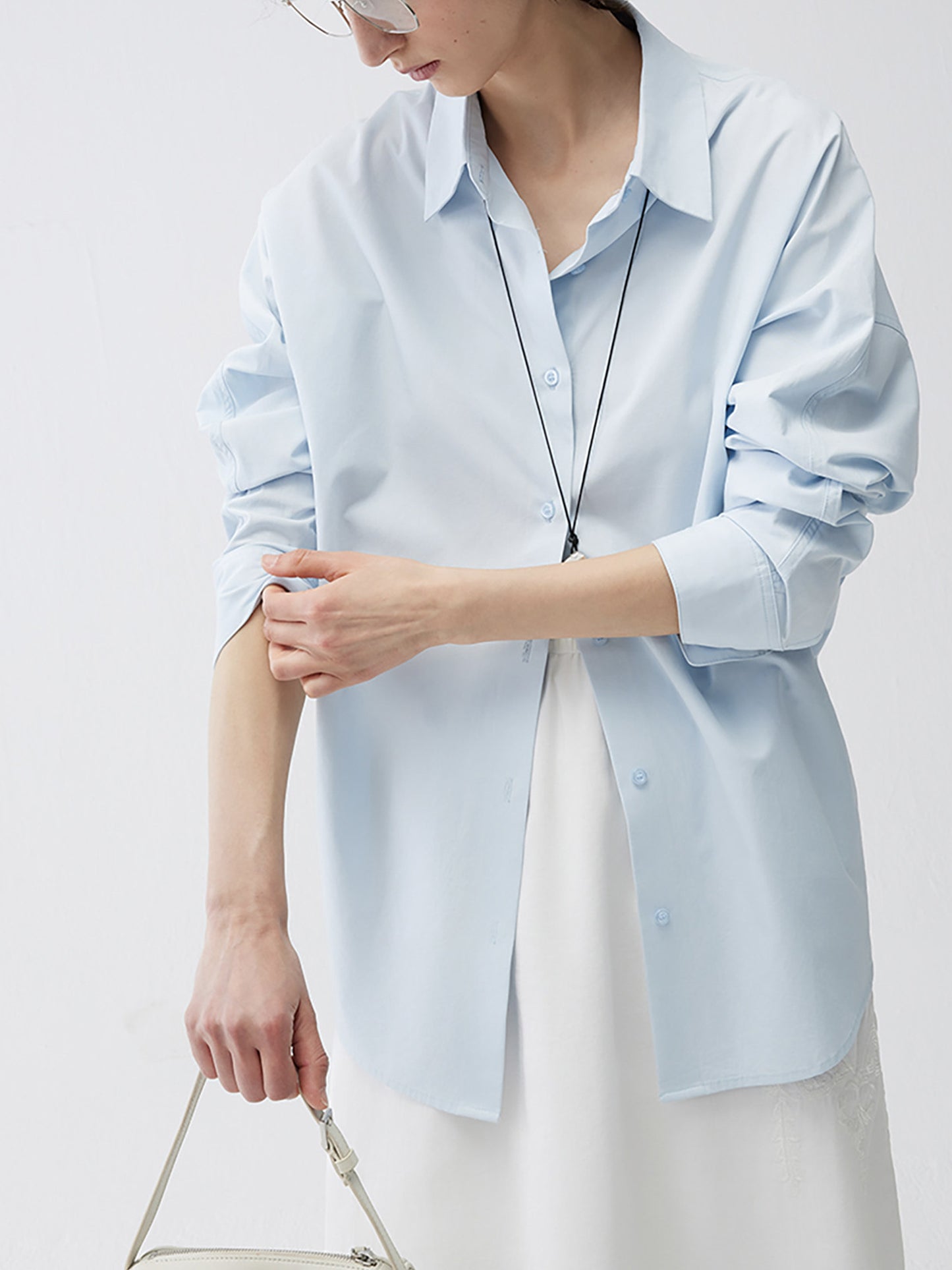 Woman wearing Arkin Studio relaxed blue tencel cotton blouse in light blue and a white skirt, with a white handbag, looking at her hand as she rolls up one sleeve with the other hand, on a white background