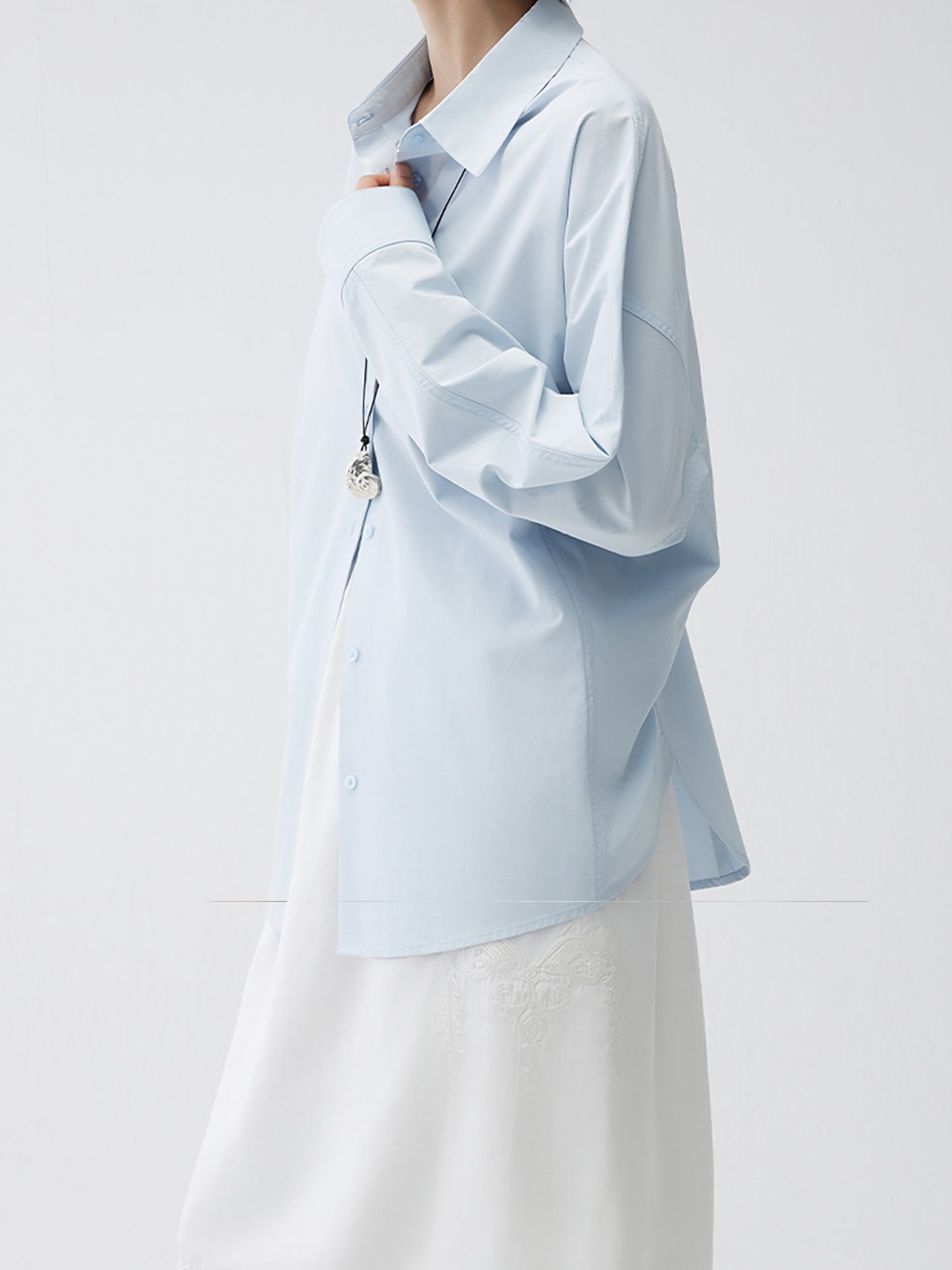 Woman wearing Arkin Studio relaxed blue tencel cotton blouse in light blue and a white skirt, standing sideways, on a white background