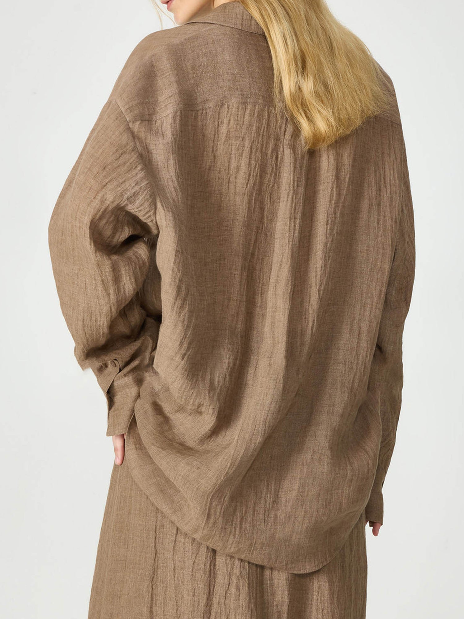 Woman with golden hair, wearing Arkin Studio relaxed crinkled linen shirt  in coffee and matching skirt, facing the white wall(back view), on a white background