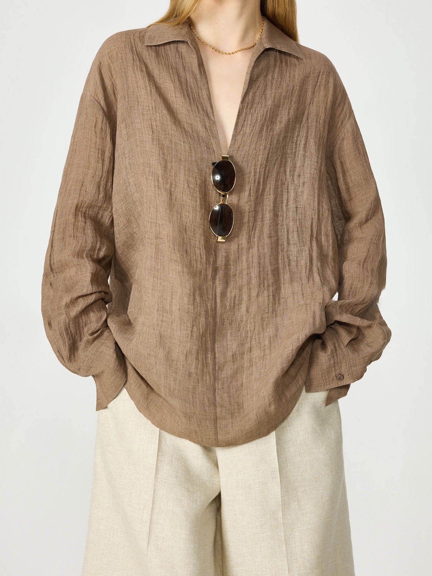 Woman wearing Arkin Studio relaxed crinkled linen shirt  in coffee and beige shorts, with a pair of glasses hanging on the shirt, hands in the shorts' pockets, standing facing the camera, on a white background