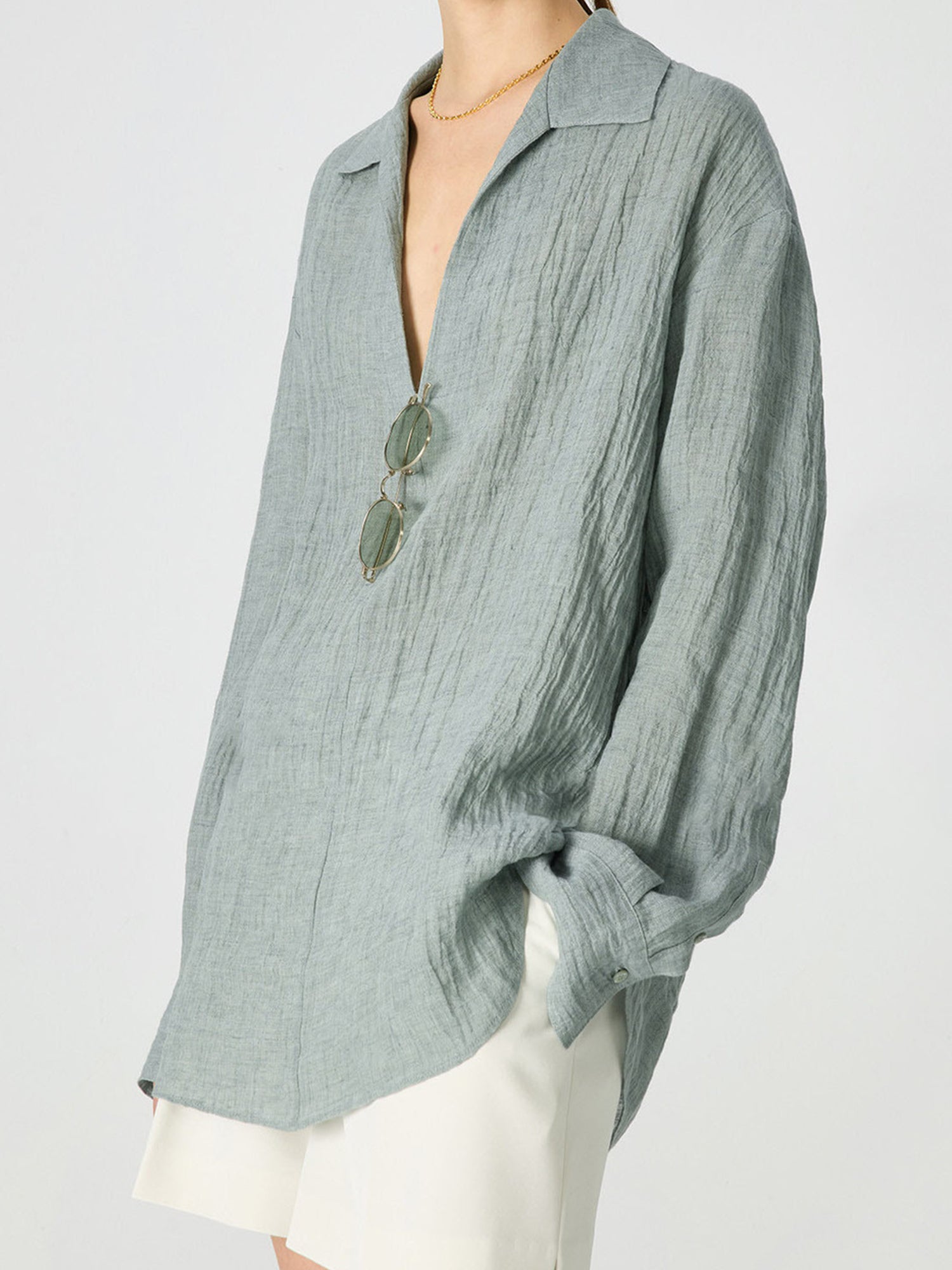 Woman wearing Arkin Studio relaxed crinkled linen shirt in mint-green and white shorts, with a pair of glasses hanging on the shirt, hands in the shorts' pockets, standing sideways, on a white background