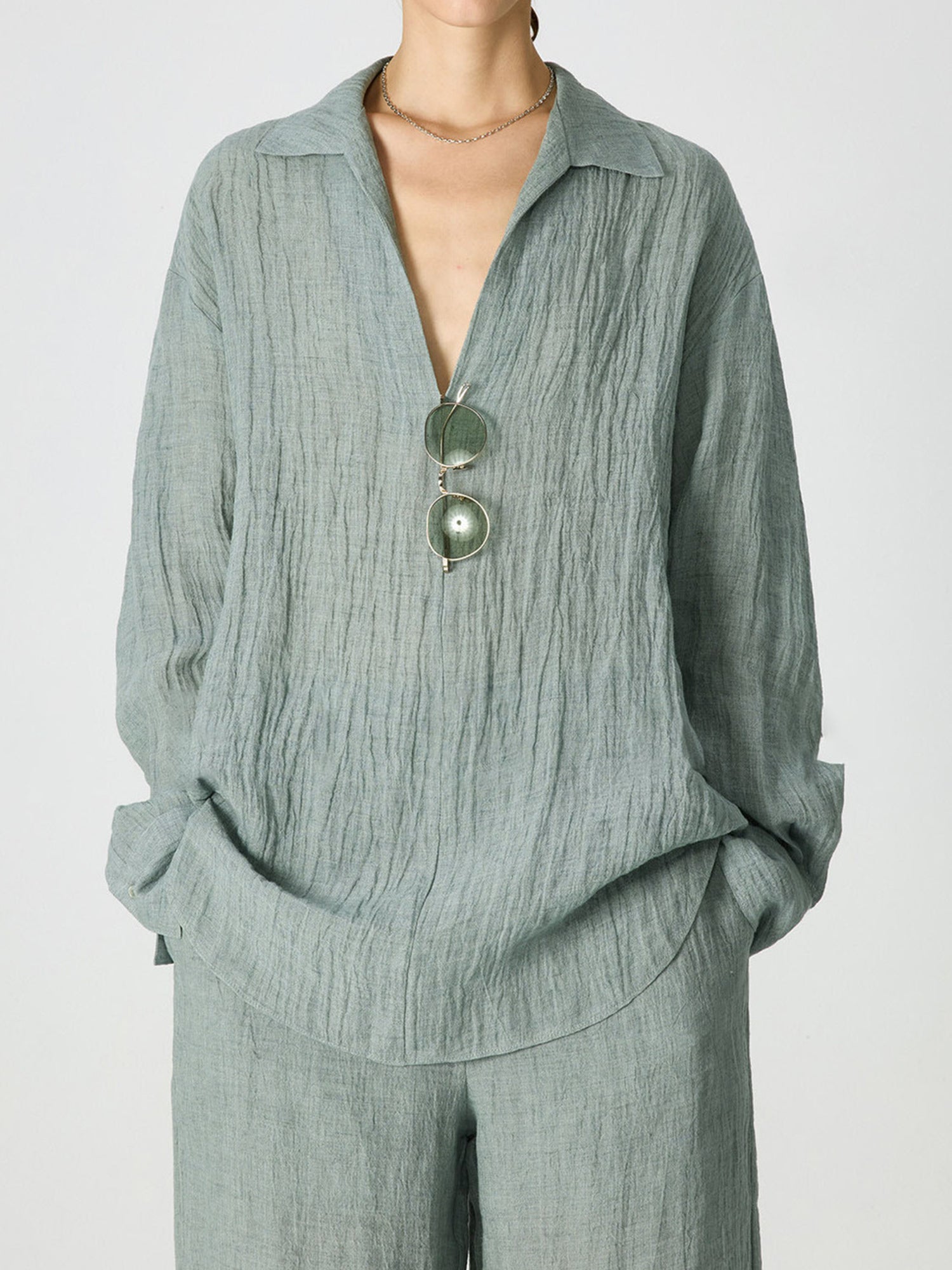 Woman wearing Arkin Studio relaxed crinkled linen shirt in mint-green and matching trousers, with a pair of glasses hanging on the shirt, hands in the trousers' pockets, standing facing the camera, on a white background