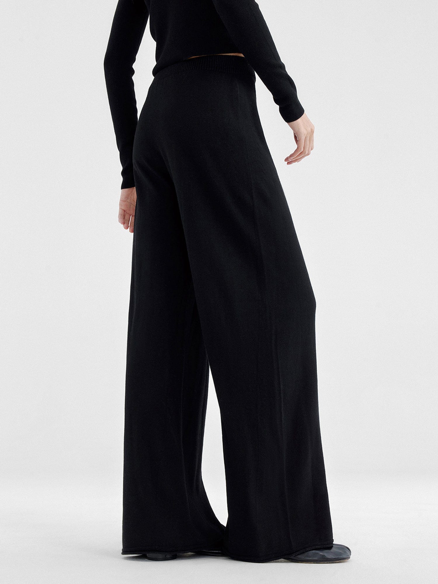 Woman wearing Arkin Studio relaxed drape knit pants in black and matching top, hands down at her sides, wearing a pair of black shoes, standing facing a white wall, on a white background