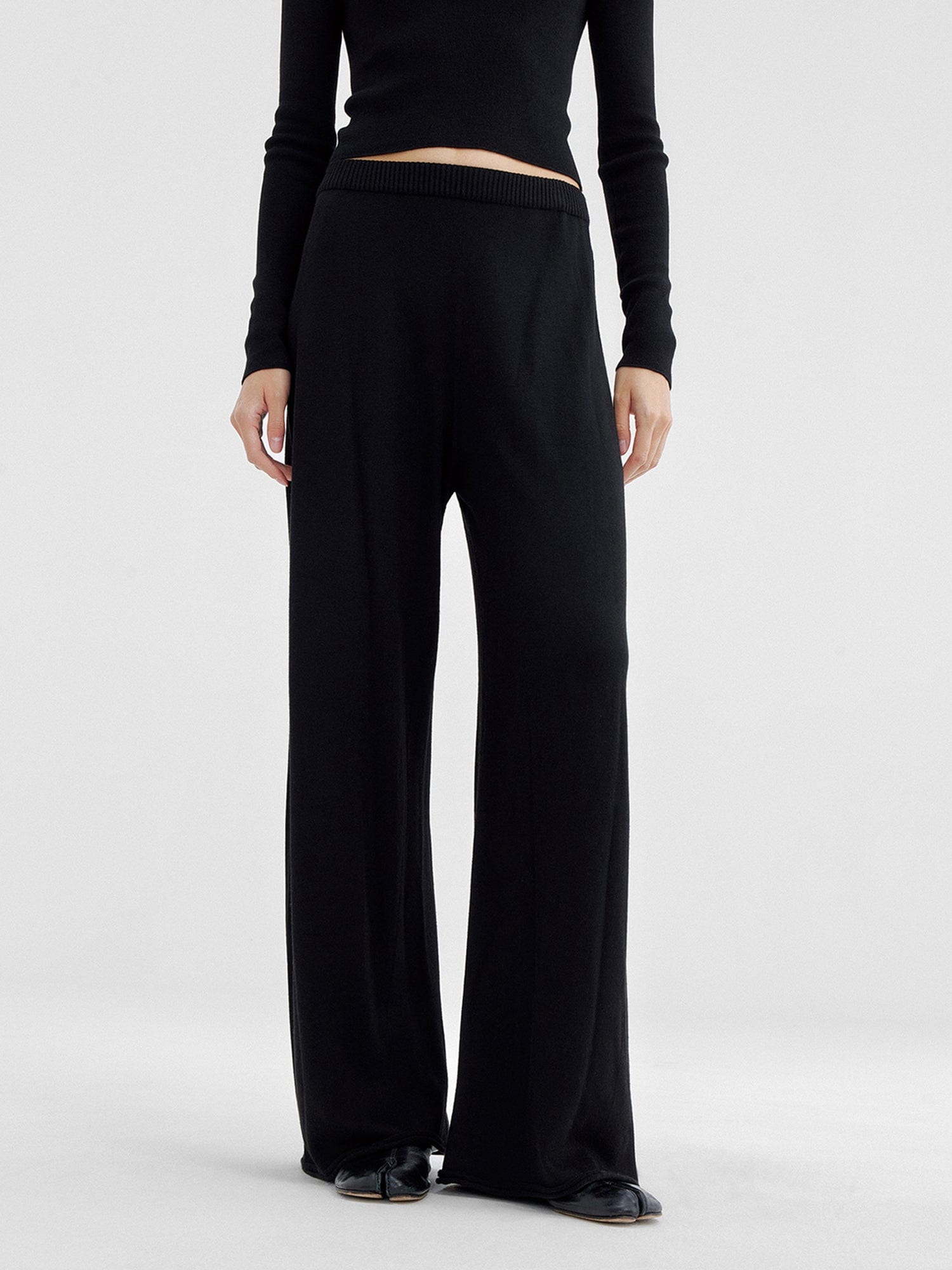Woman wearing Arkin Studio relaxed drape knit pants in black and matching top, hands down at her sides, wearing a pair of black shoes, standing facing the camera, on a white background