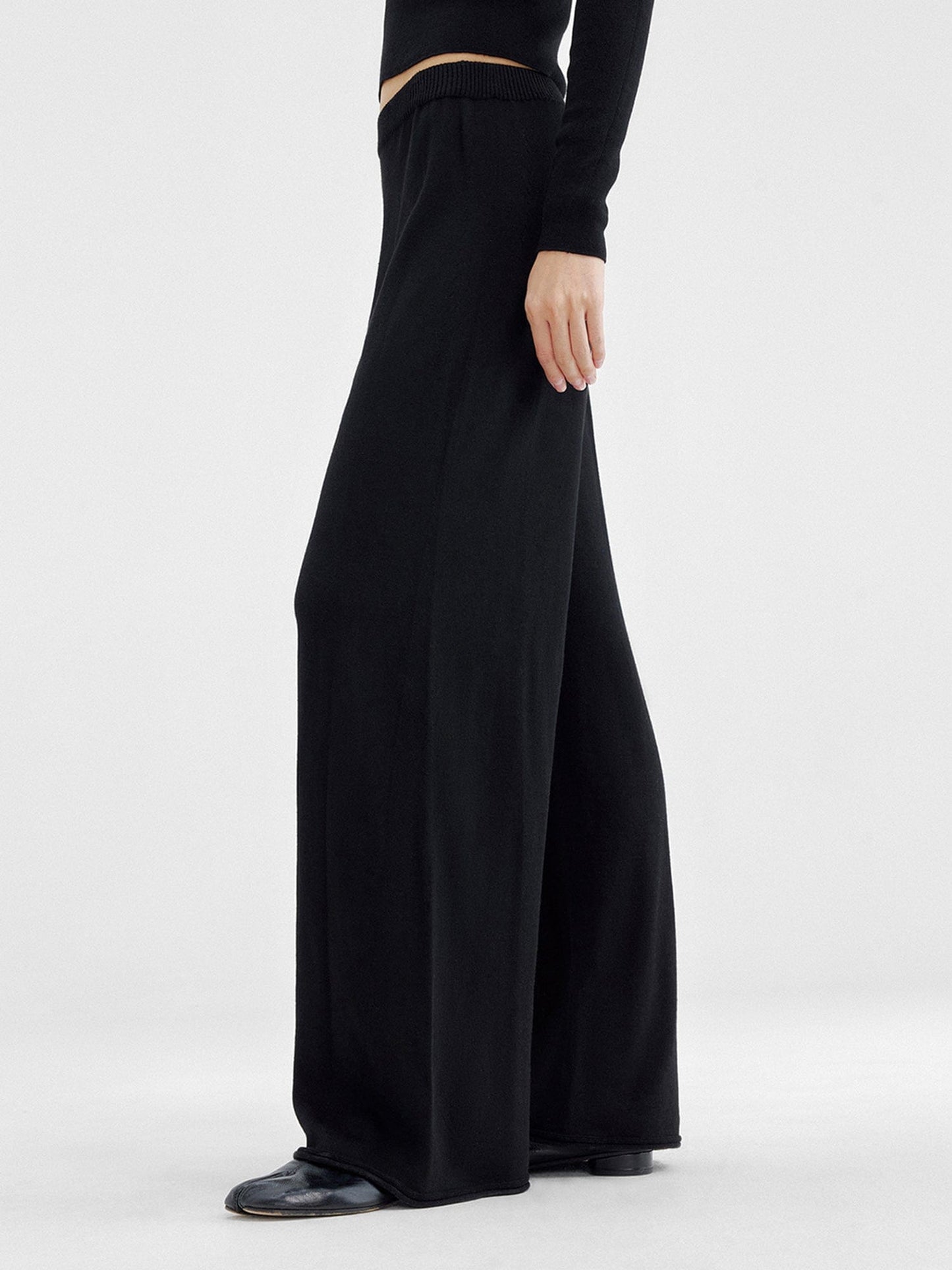 Woman wearing Arkin Studio relaxed drape knit pants in black and matching top, hands down at her sides, wearing a pair of black shoes, standing sideways, on a white background