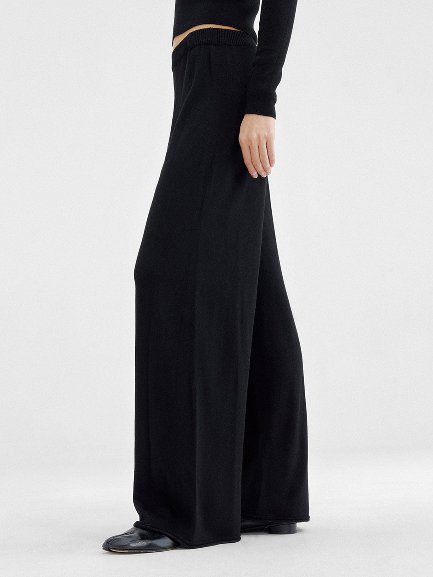 Woman wearing Arkin Studio relaxed drape knit pants in black and matching top, hands down at her sides, wearing a pair of black shoes, standing sideways, on a white background
