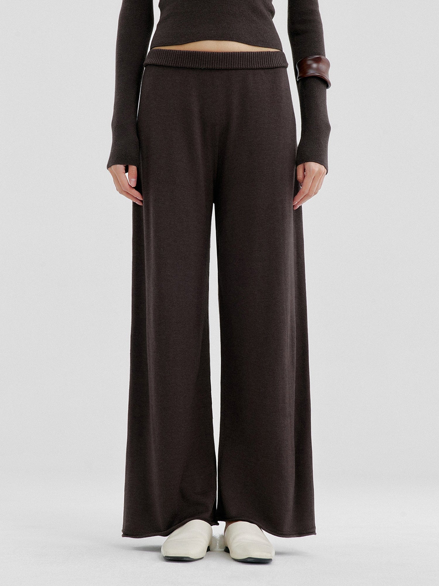 Woman wearing Arkin Studio relaxed drape knit pants in dark brown and matching top, hands down at her sides, wearing a pair of white shoes, standing facing the camera, on a white background