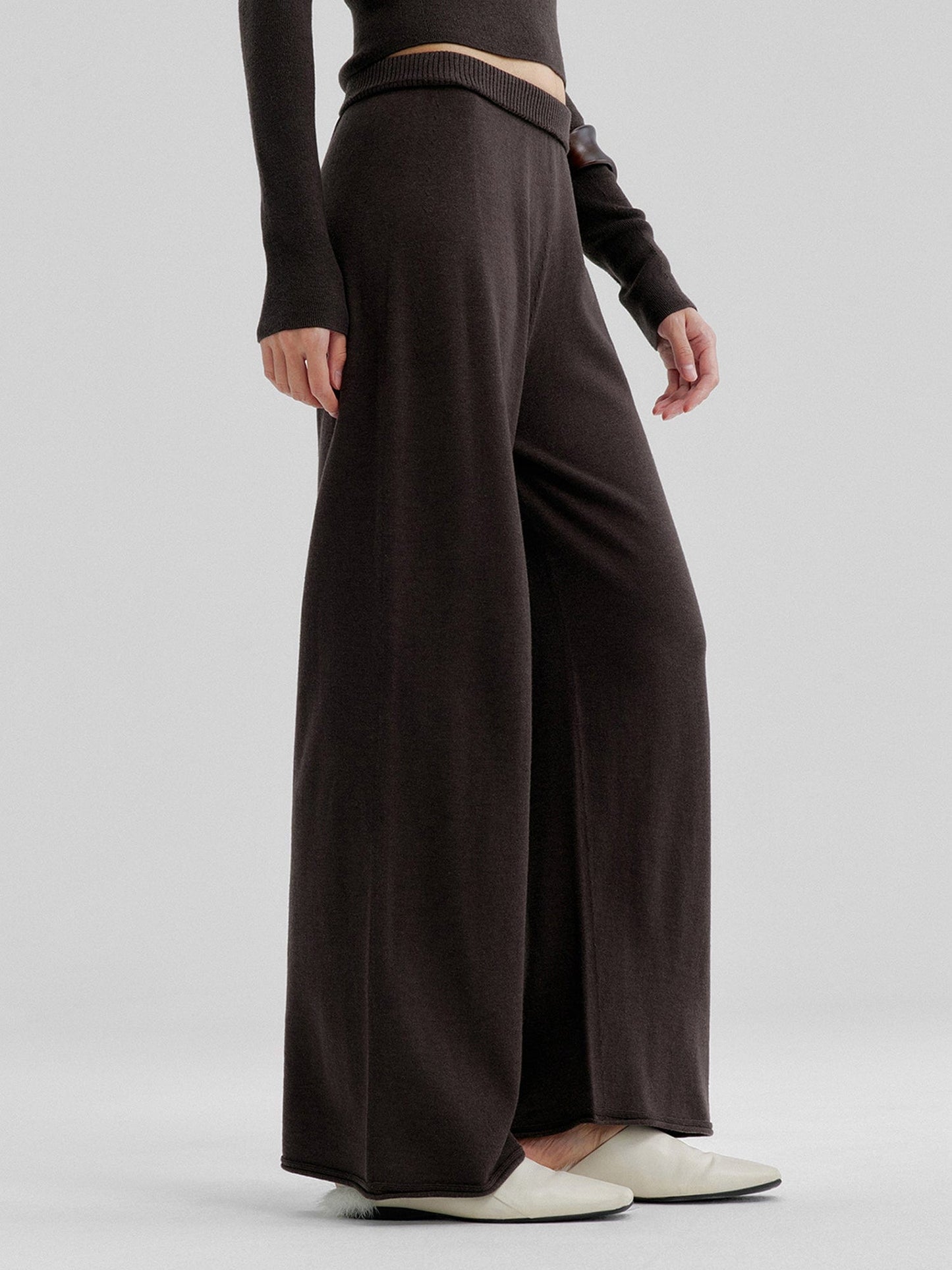 Woman wearing Arkin Studio relaxed drape knit pants in dark brown and matching top, hands down at her sides, wearing a pair of white shoes, standing sideways, on a white background
