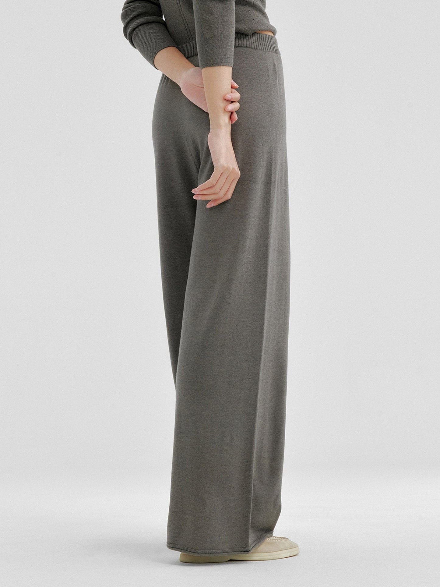 Woman wearing Arkin Studio relaxed drape knit pants in gray and matching top, hands down behind her back, wearing a pair of beige shoes, standing facing a white wall, on a white background