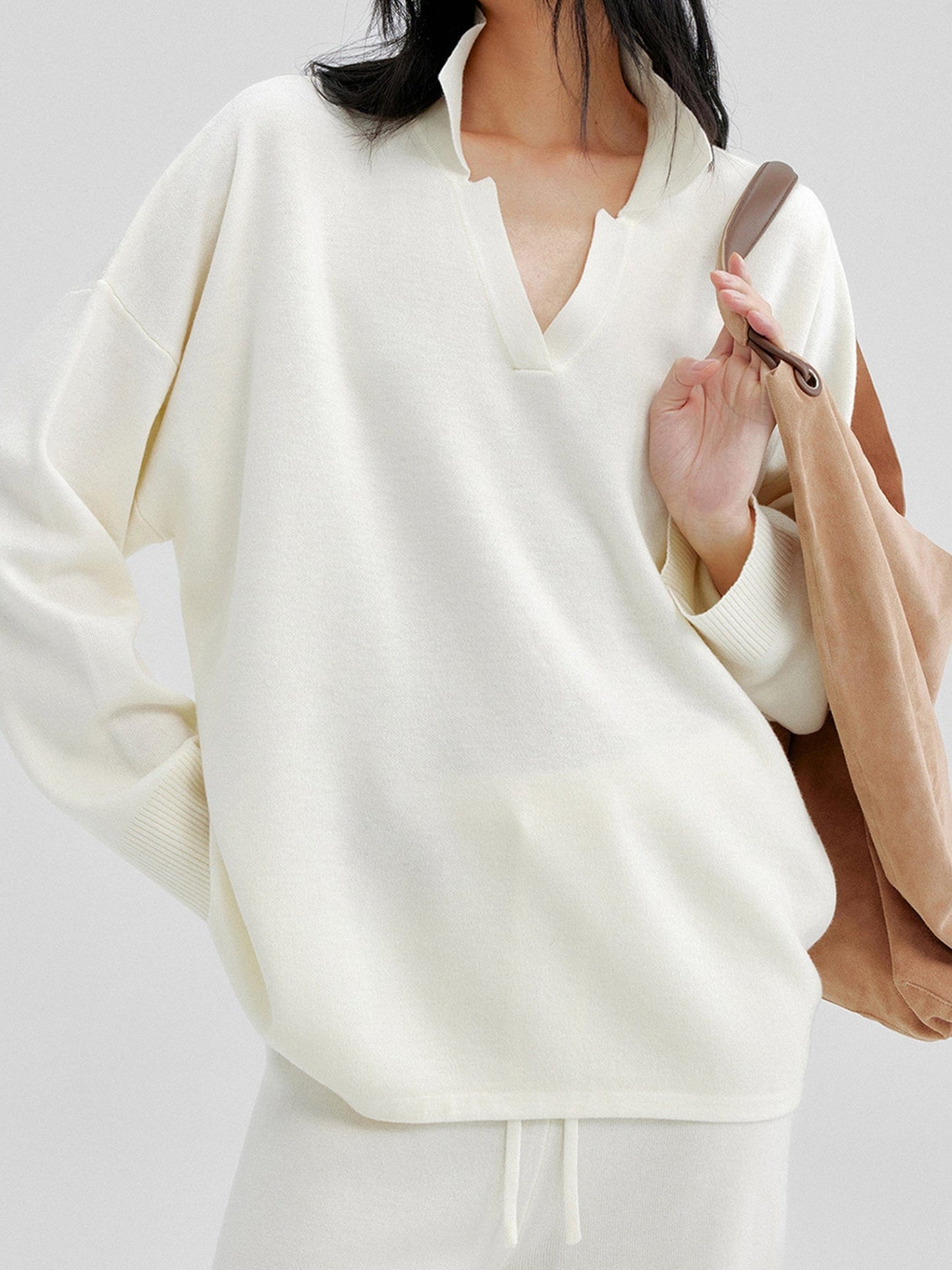 Close up of a woman wearing Arkin Studio relaxed v-neck wool blend sweater in ivory and matching pants, a brown bag on her left shoulder, focus on the upper part of her, standing facing the camera, on a white background