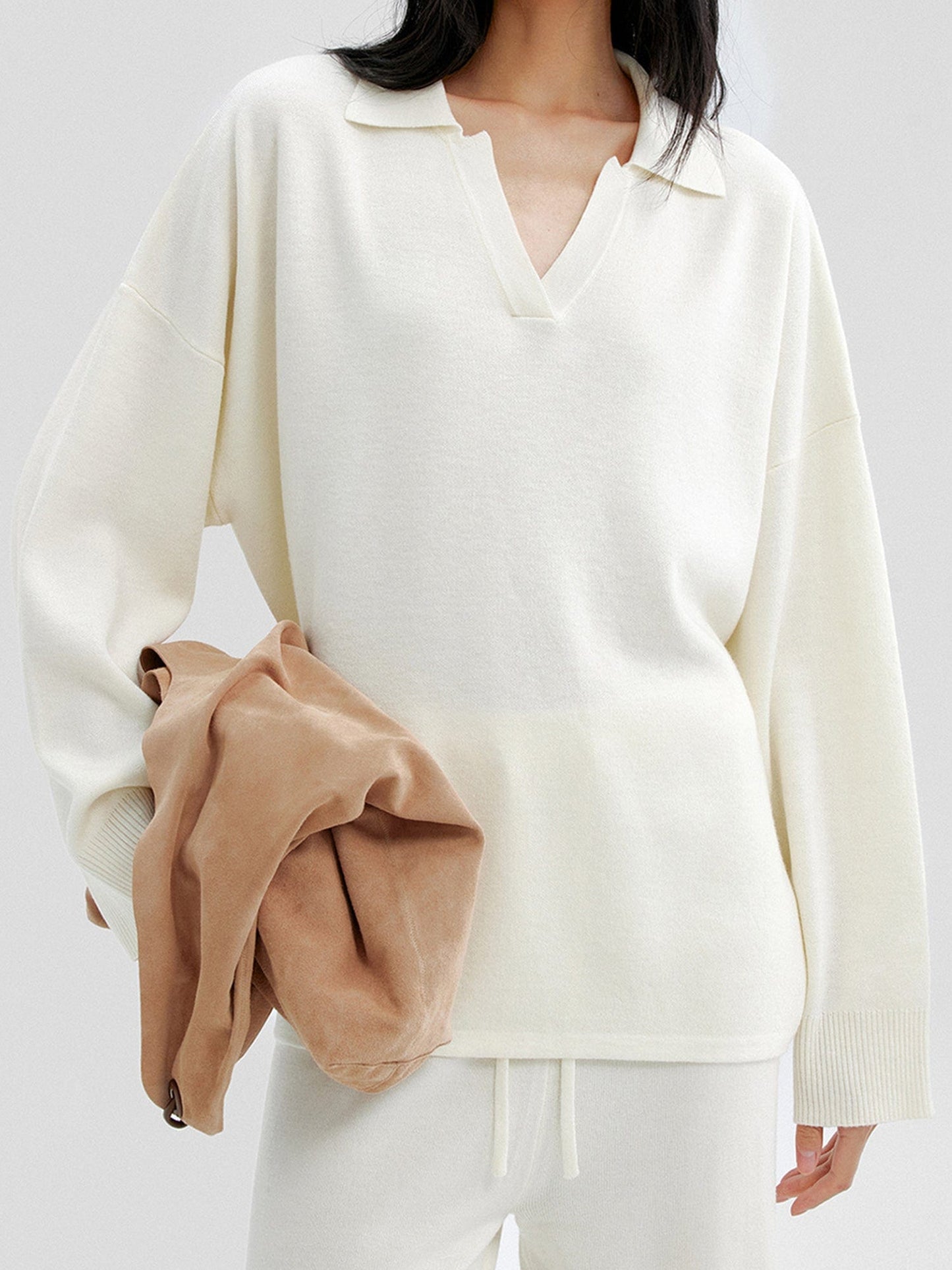 Woman wearing Arkin Studio relaxed v-neck wool blend sweater in ivory and matching pants, holding a brown bag, standing facing the camera, on a white background
