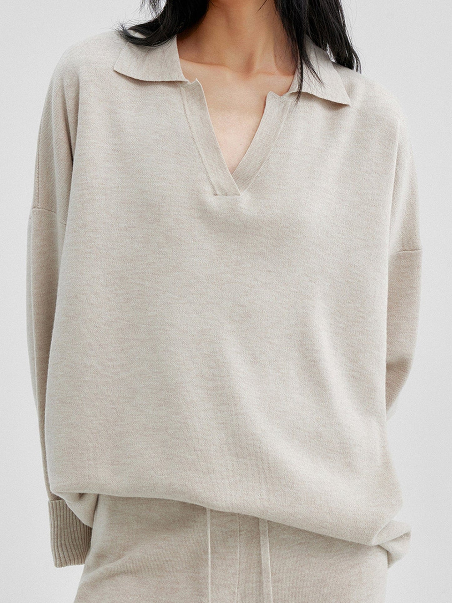 Close up of a woman wearing Arkin Studio relaxed v-neck wool blend sweater in taupe and matching pants, hands behind her back, focus on the upper part of her, standing facing the camera, on a white background