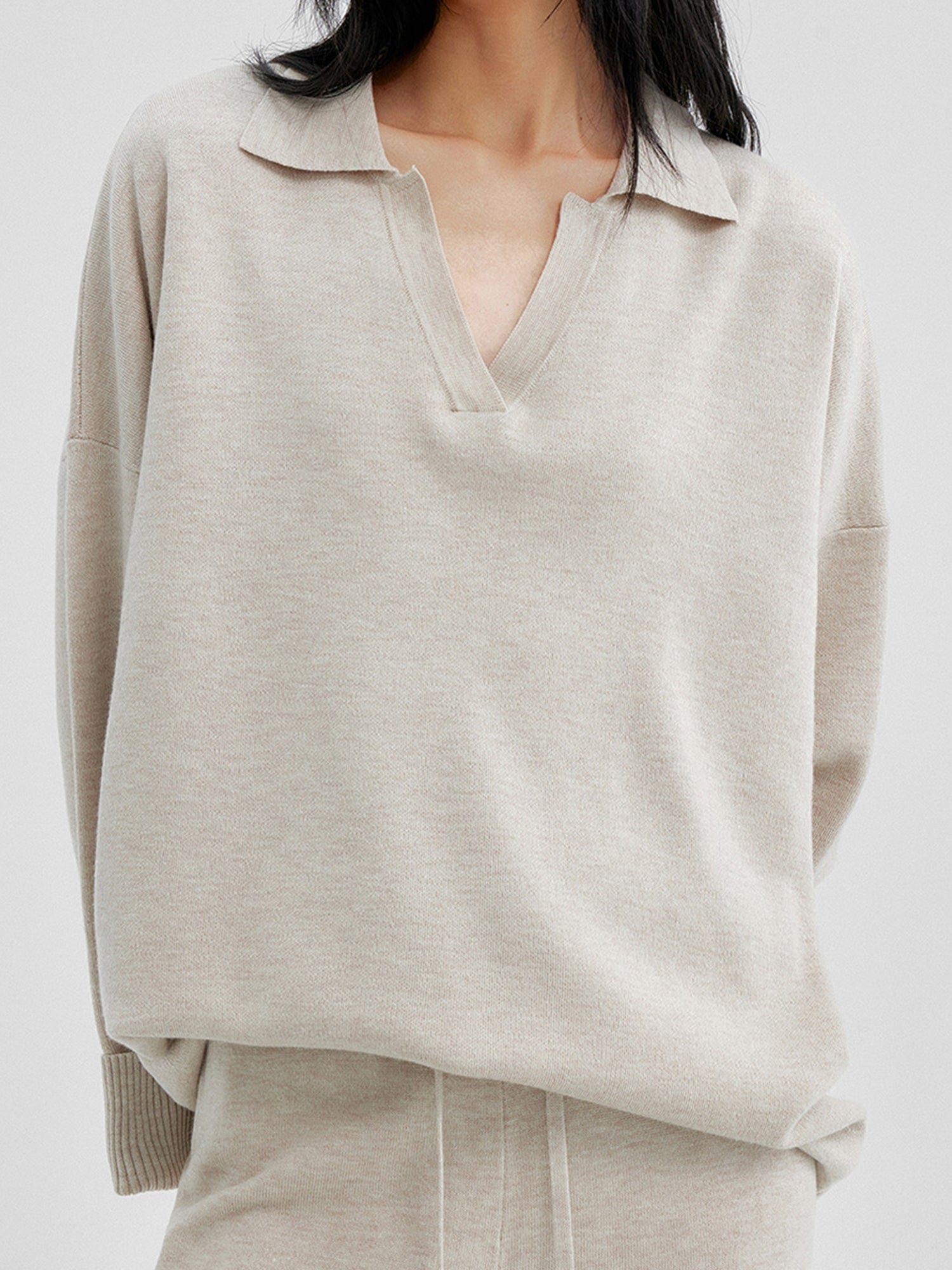 Close up of a woman wearing Arkin Studio relaxed v-neck wool blend sweater in taupe and matching pants, hands behind her back, focus on the upper part of her, standing facing the camera, on a white background