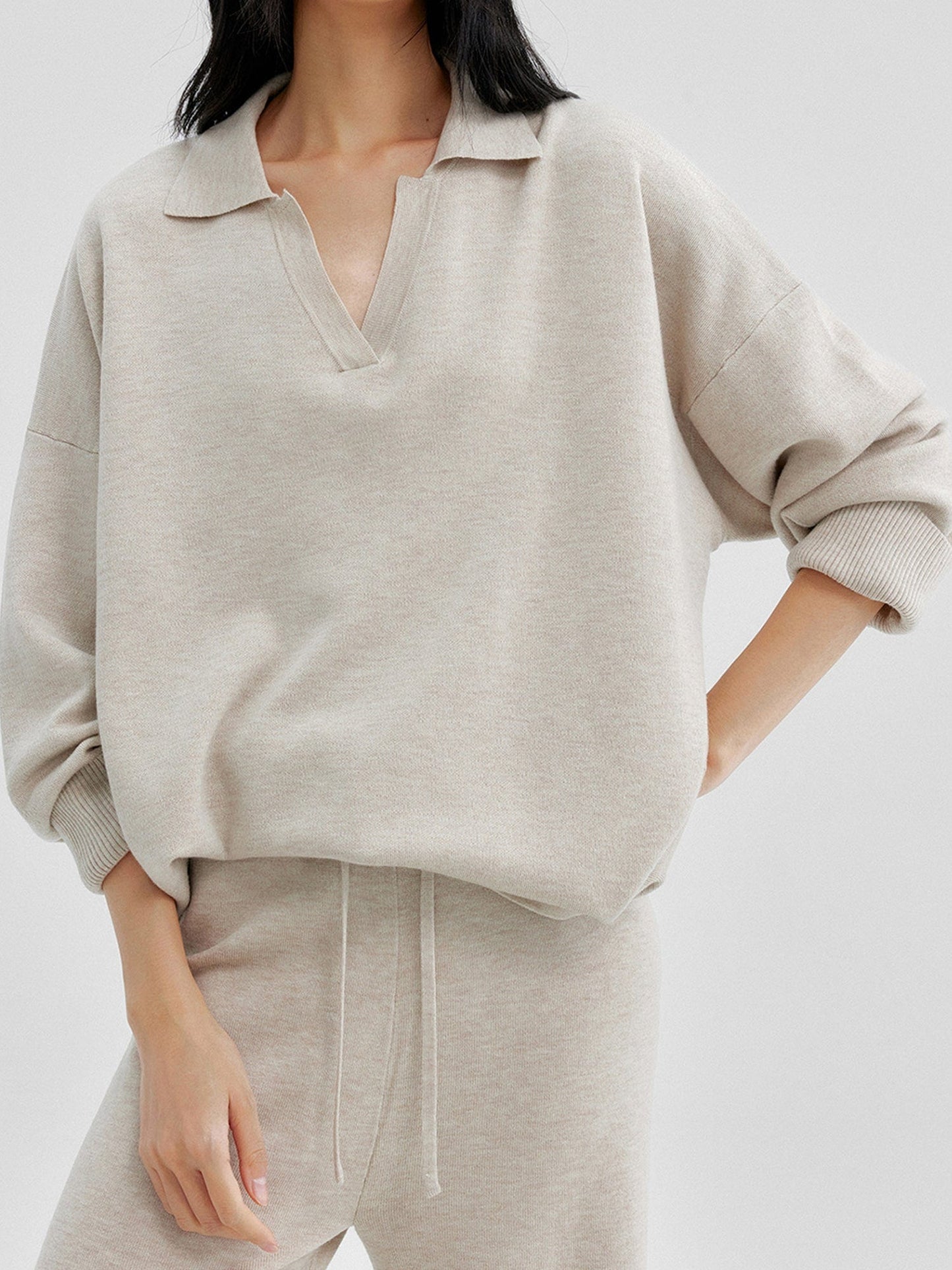 Woman wearing Arkin Studio relaxed v-neck wool blend sweater in taupe and matching pants, one hand behind her back and another on her leg, standing facing the camera, on a white background