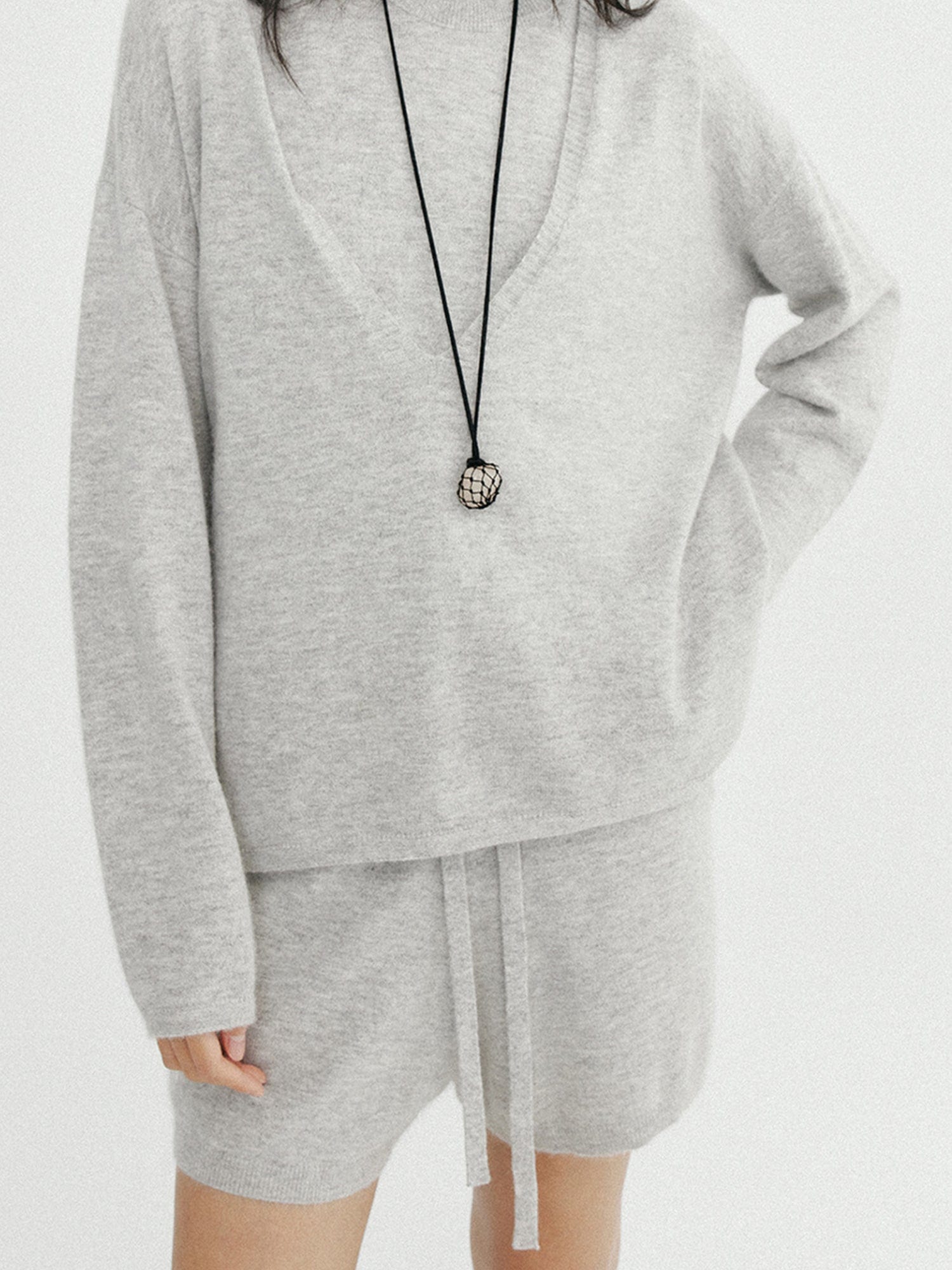 Woman wearing Arkin Studio ribbed knit drawstring shorts in gray and matching top, one hand in a pocket, standing facing the camera, with a long pendant hanging on her chest, on a white background
