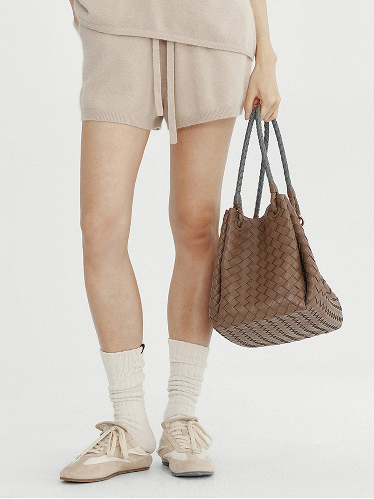 Woman wearing Arkin Studio ribbed knit drawstring shorts in taupe and matching top, holding a brown bag, standing facing the camera, on a white background