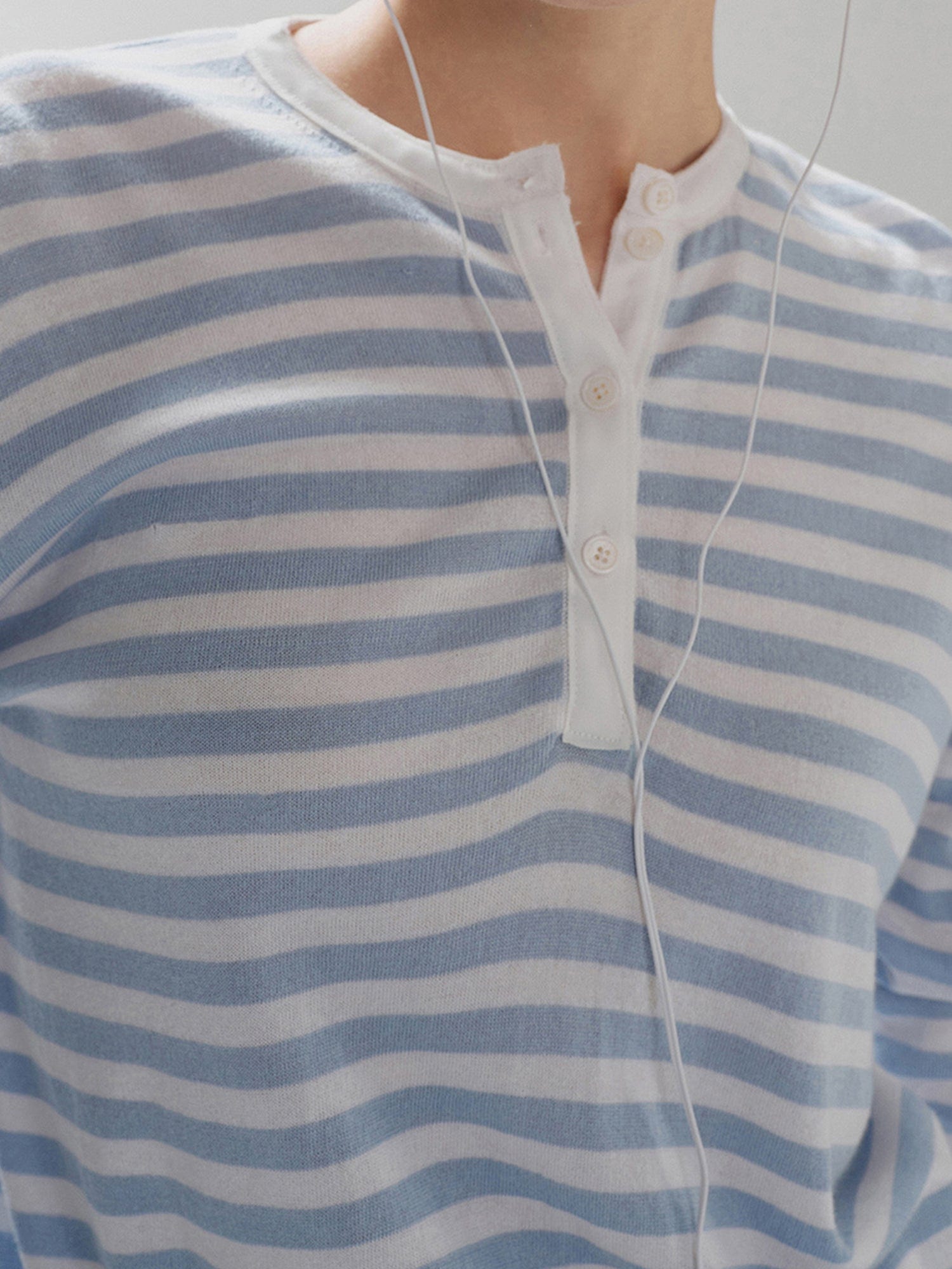 Close-up of a woman's chest wearing Arkin Studio light blue striped henley long sleeve tee, with headphone cord draped across her chest, standing facing the camera, on a white background