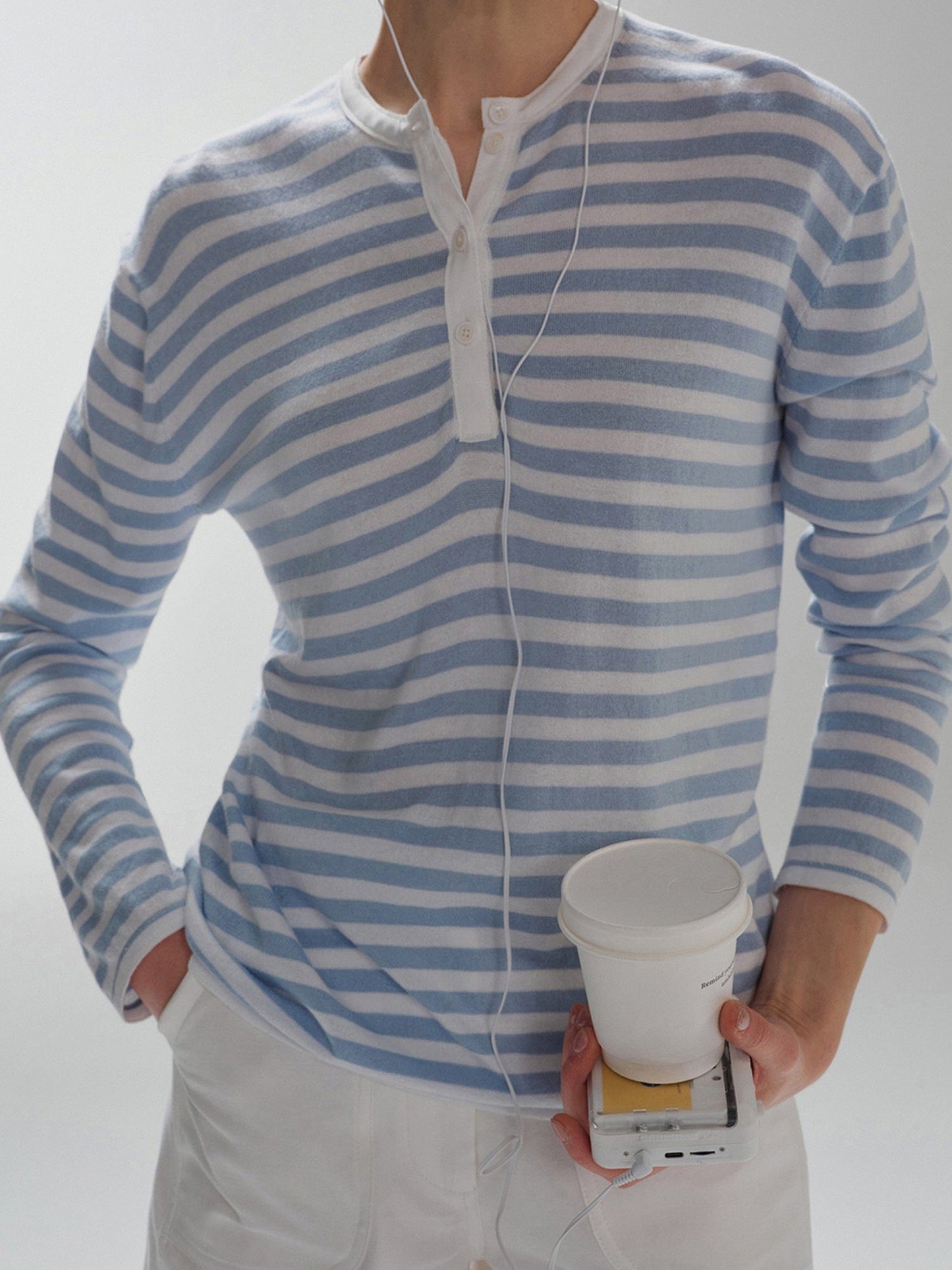 Woman wearing Arkin Studio Striped Henley Long Sleeve Tee in Light Blue, with a paper cup in her hand, standing facing the camera, on a white background
