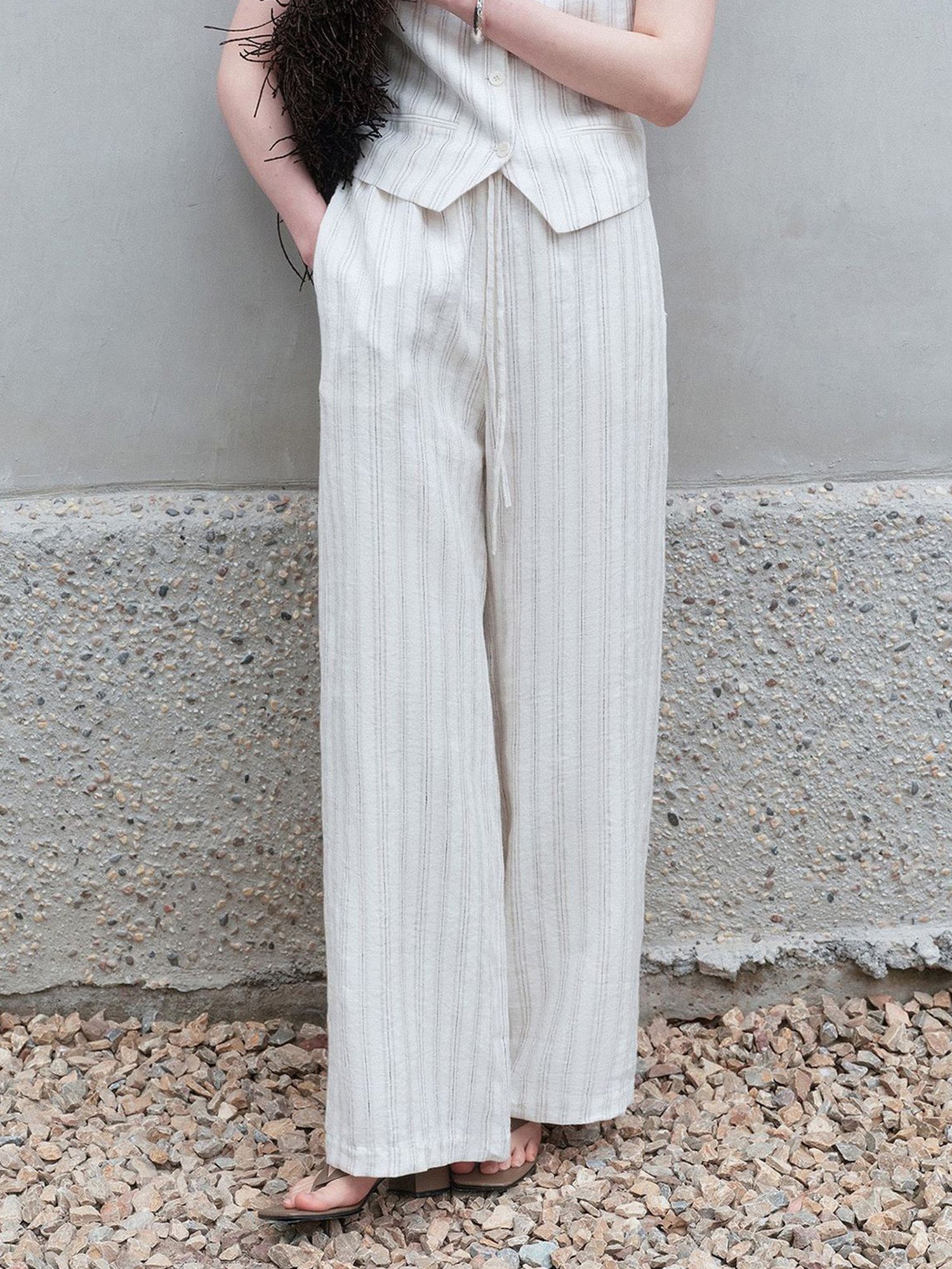 Woman wearing Arkin Studio striped linen drawstring wide leg pants in ivory and matching vest, with a brown flat bag tucked under her arm, standing on a stone floor, facing the camera