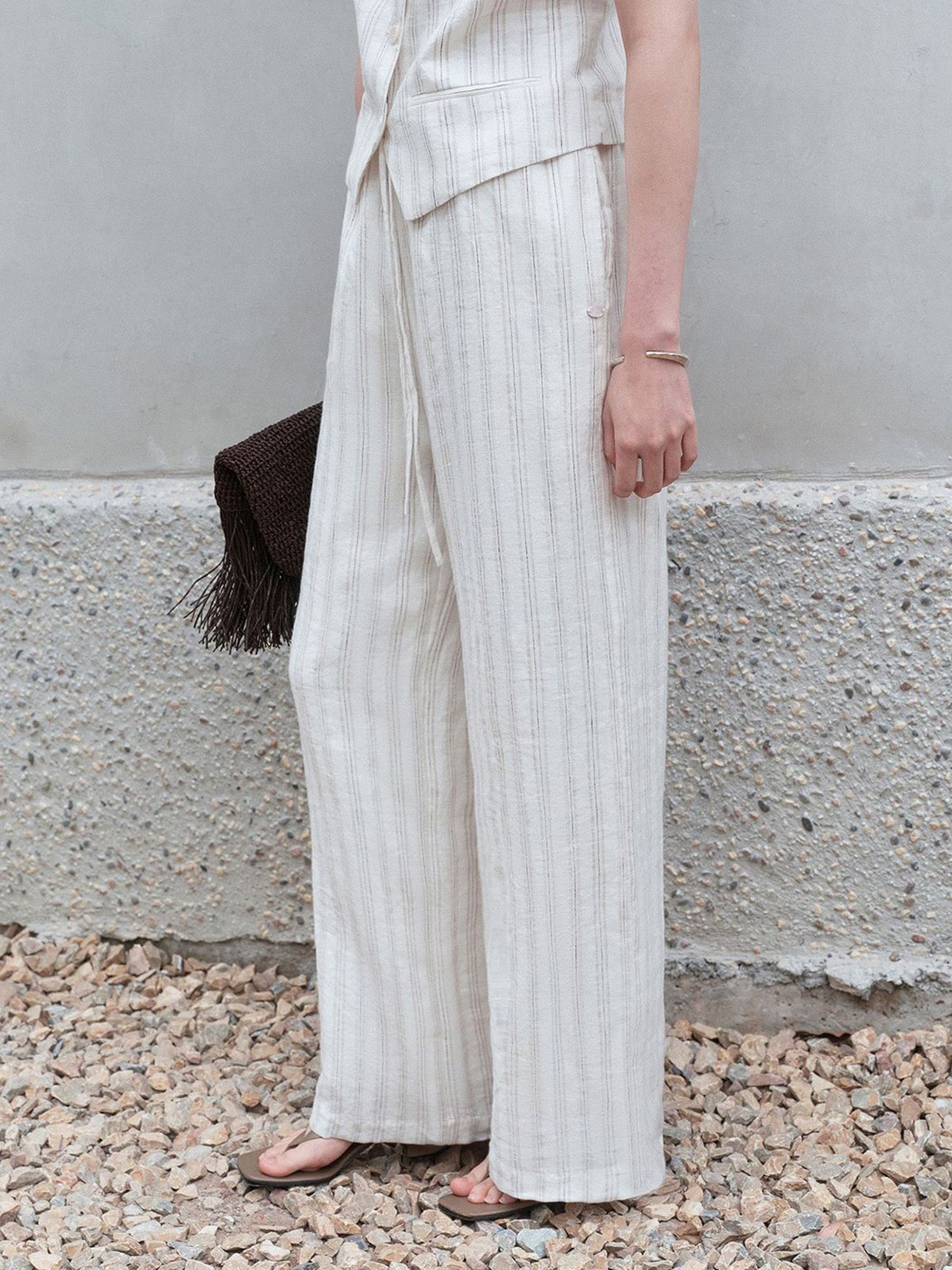 Woman wearing Arkin Studio striped linen drawstring wide leg pants in ivory and matching vest, holding a brown flat bag, standing sideways on a stone floor