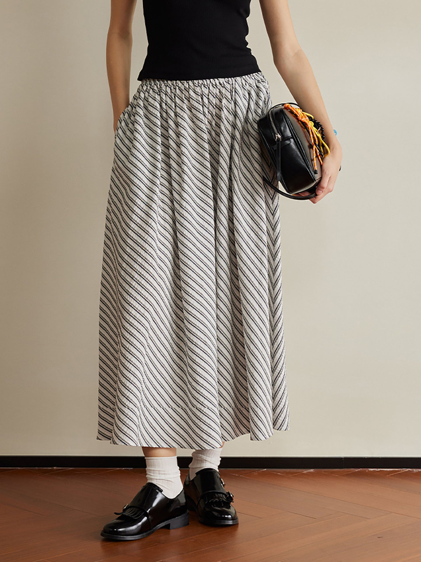 Woman wearing Arkin Studio striped pleated a-line midi skirt and a black top, wearing a pair of black loafers, holding a black handbag, standing facing the camera on the brown floor, a white wall behind her