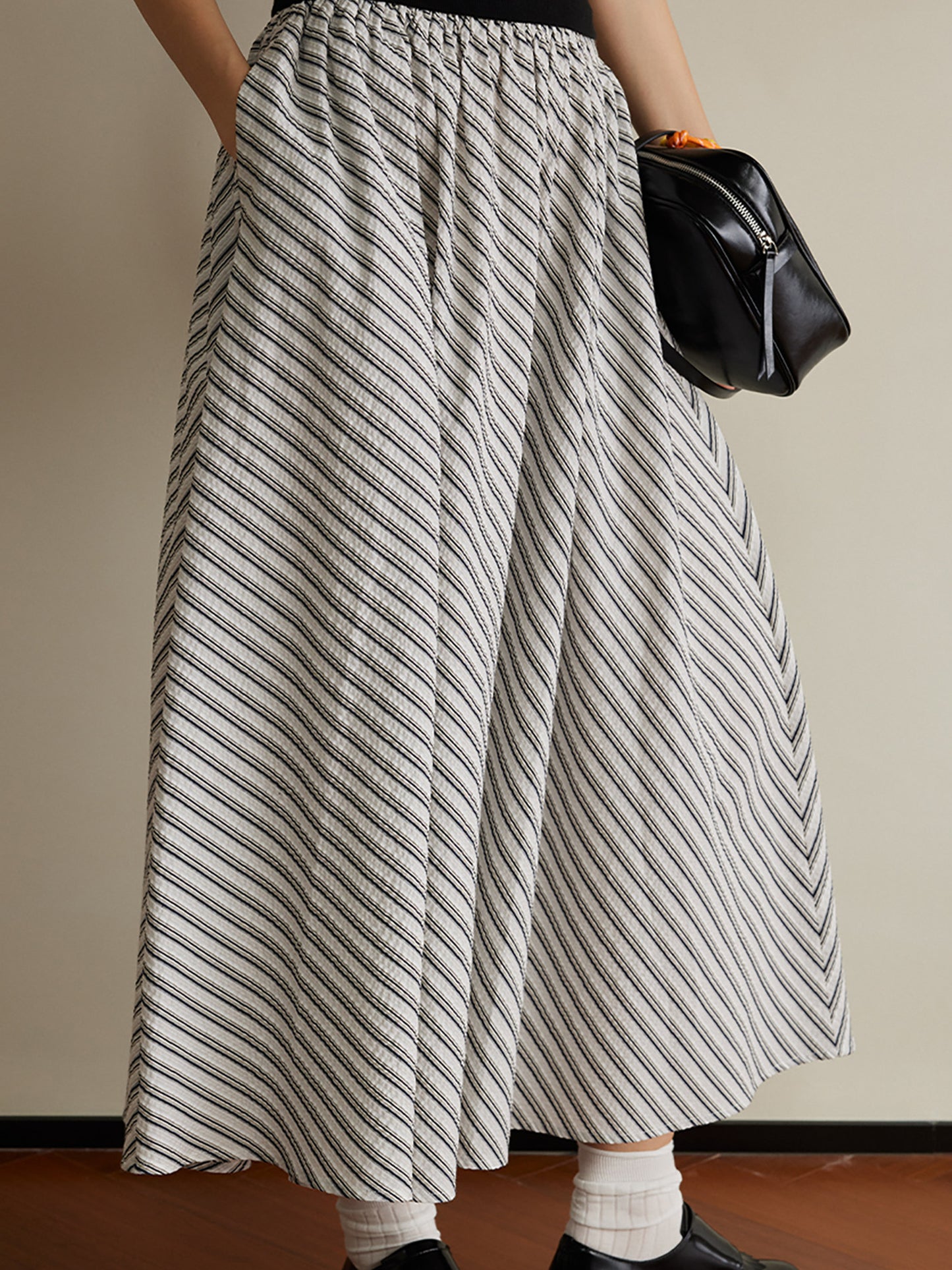 Woman wearing Arkin Studio striped pleated a-line midi skirt, wearing a pair of black loafers, holding a black handbag, standing facing the camera on the brown floor, a white wall behind her