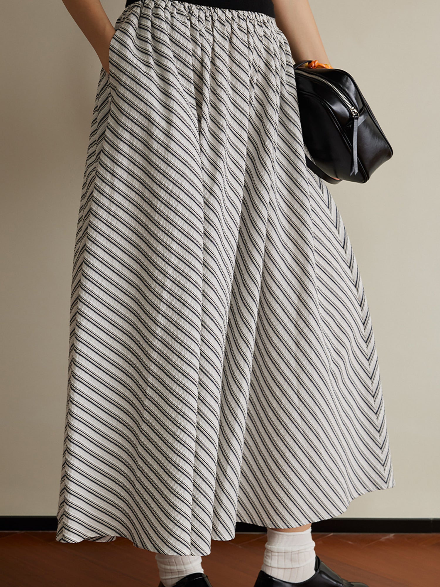 Woman wearing Arkin Studio striped pleated a-line midi skirt, wearing a pair of black loafers, holding a black handbag, standing facing the camera on the brown floor, a white wall behind her