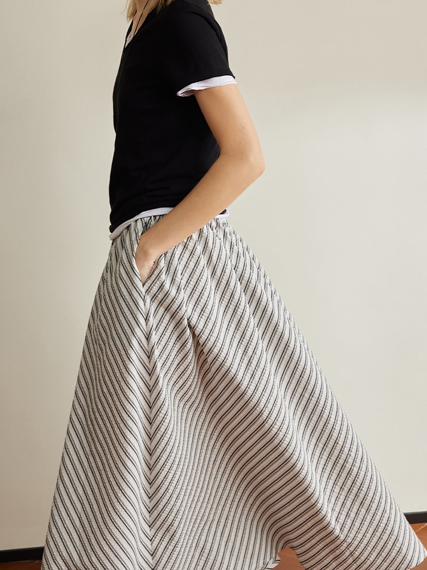 Woman wearing Arkin Studio striped pleated a-line midi skirt and a black top, walking in side view with one hand in the skirt pocket, a white wall behind her