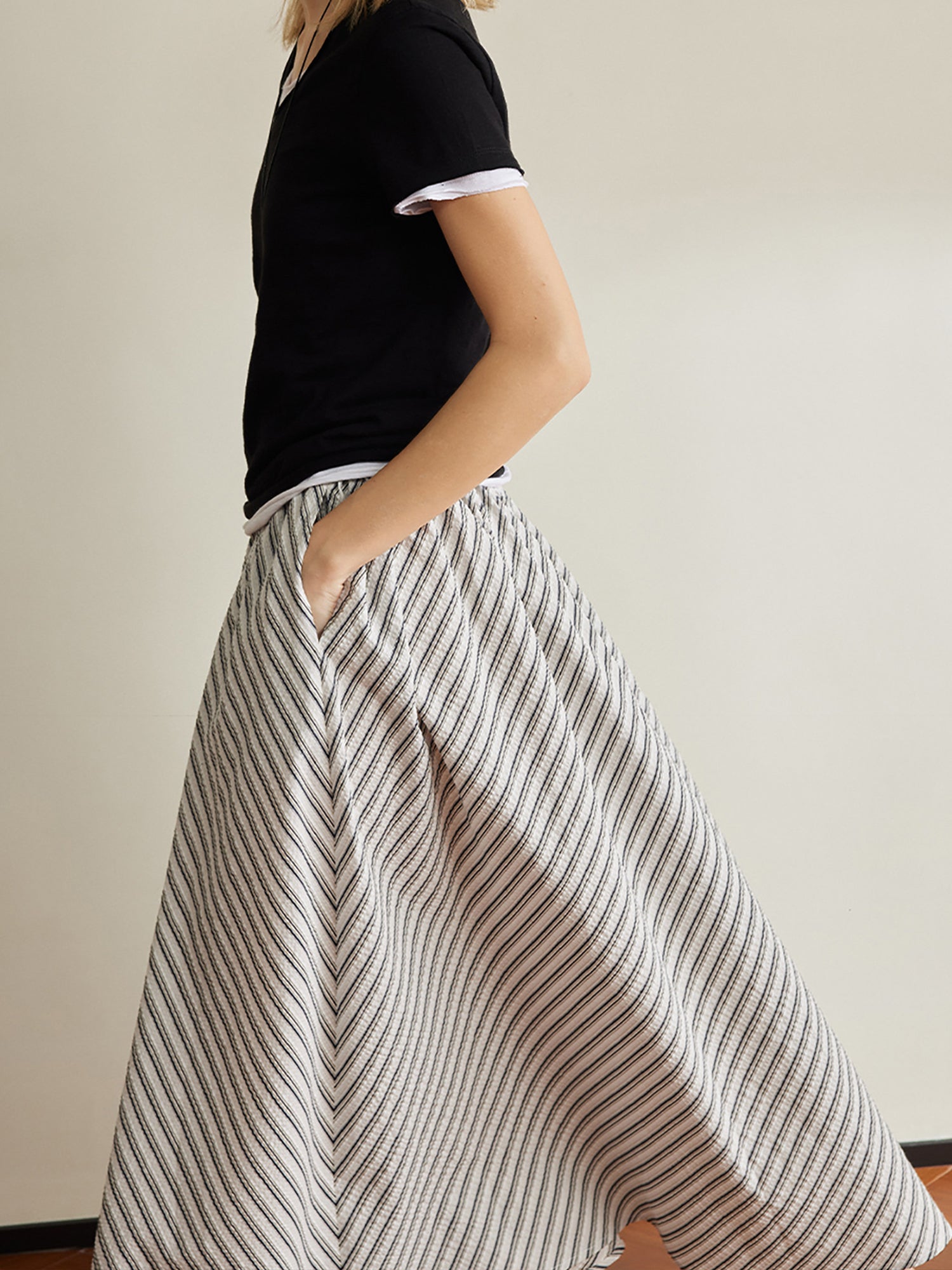 Woman wearing Arkin Studio striped pleated a-line midi skirt and a black top, walking in side view with one hand in the skirt pocket, a white wall behind her