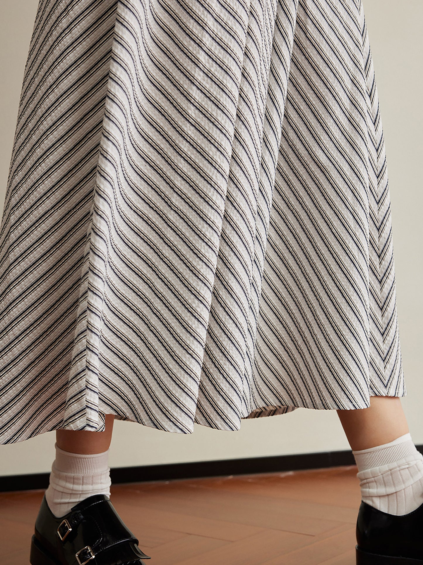 Woman wearing Arkin Studio striped pleated a-line midi skirt, walking with white socks and black loafers on the brown floor, a white wall behind her