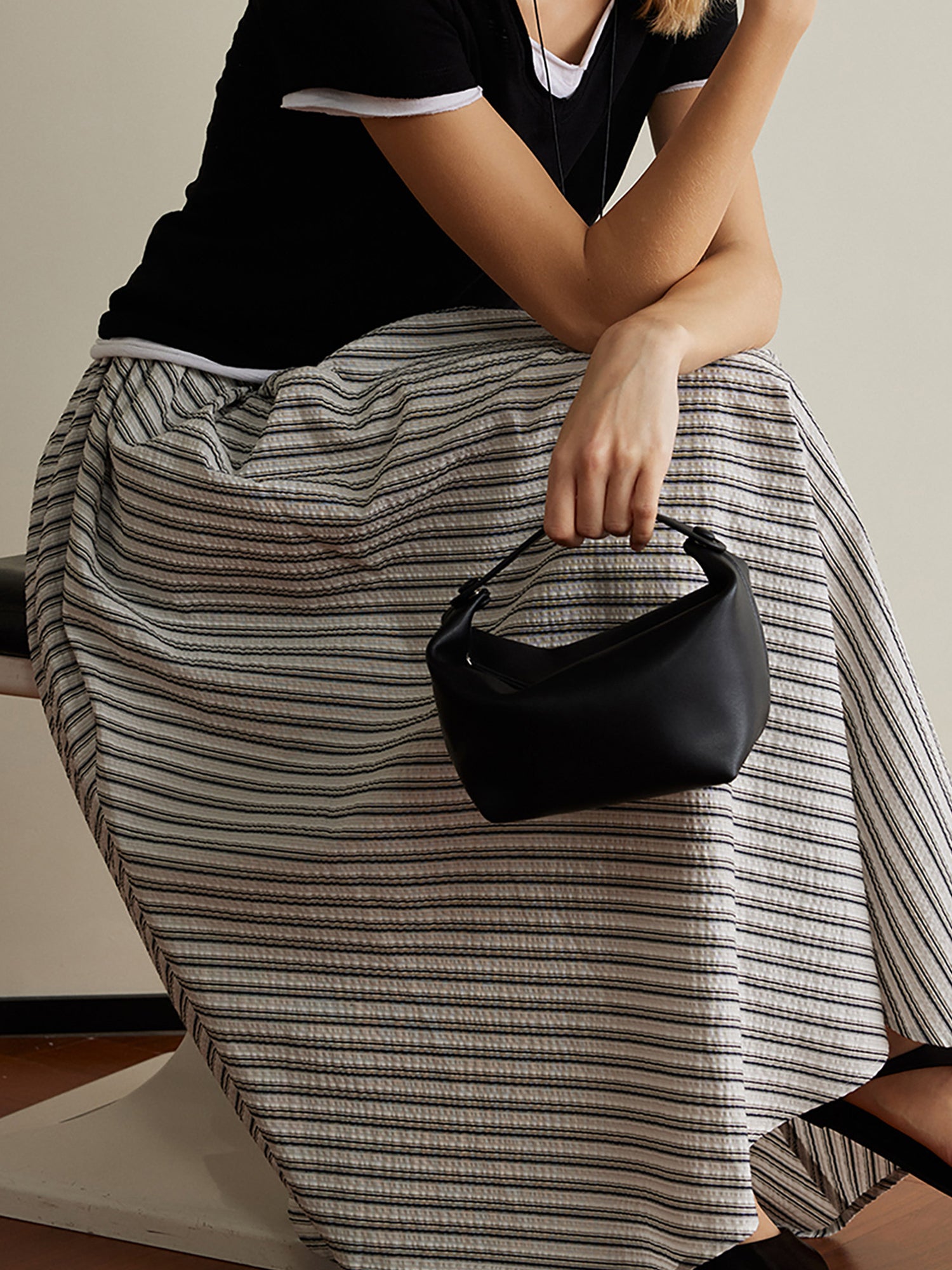 Woman wearing Arkin Studio striped pleated a-line midi skirt, sitting on a stool with a three-quarter turn on the brown floor, holding a black handbag, a white wall behind her